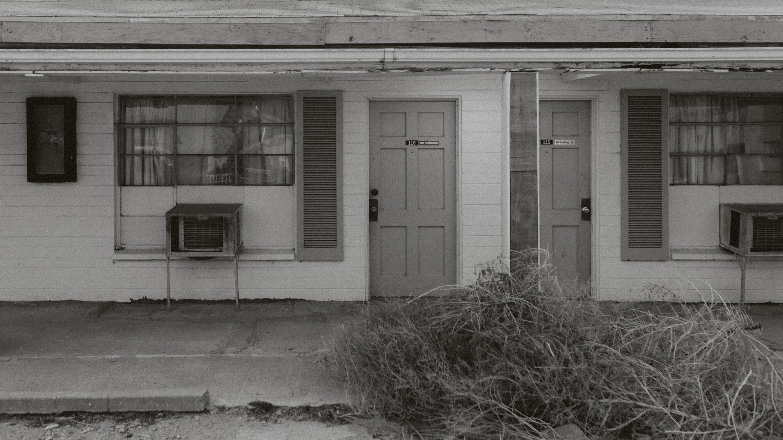 Façade d'un bâtiment résidentiel en bois avec deux portes et deux fenêtres, un climatiseur à chaque côté, et un tas de branches mortes devant. Style en noir et blanc.