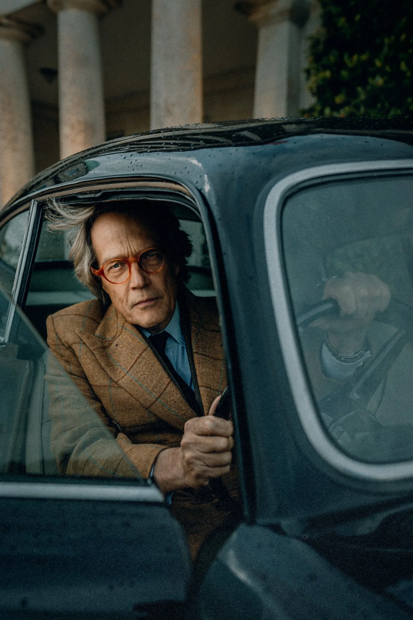 Un homme âgé avec des cheveux gris, portant des lunettes rondes et un blazer marron, regarde par la fenêtre d'une voiture. Il tient la porte de la voiture ouverte avec sa main.