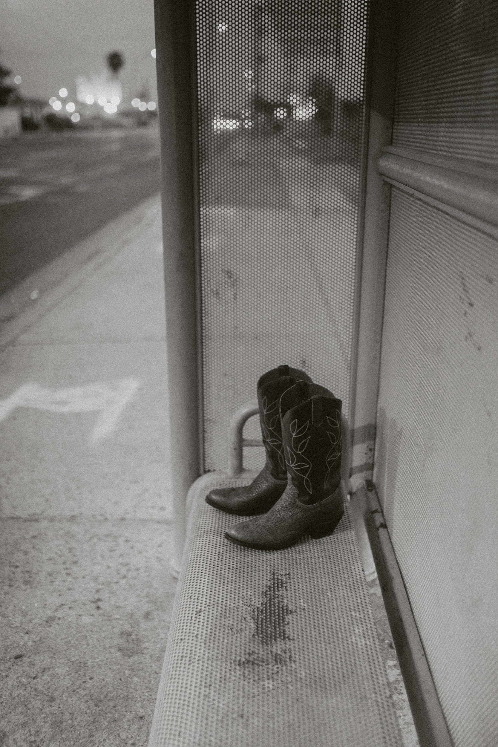Une paire de bottes de cow-boy posée sur un banc dans un arrêt de bus, l'image est en noir et blanc.