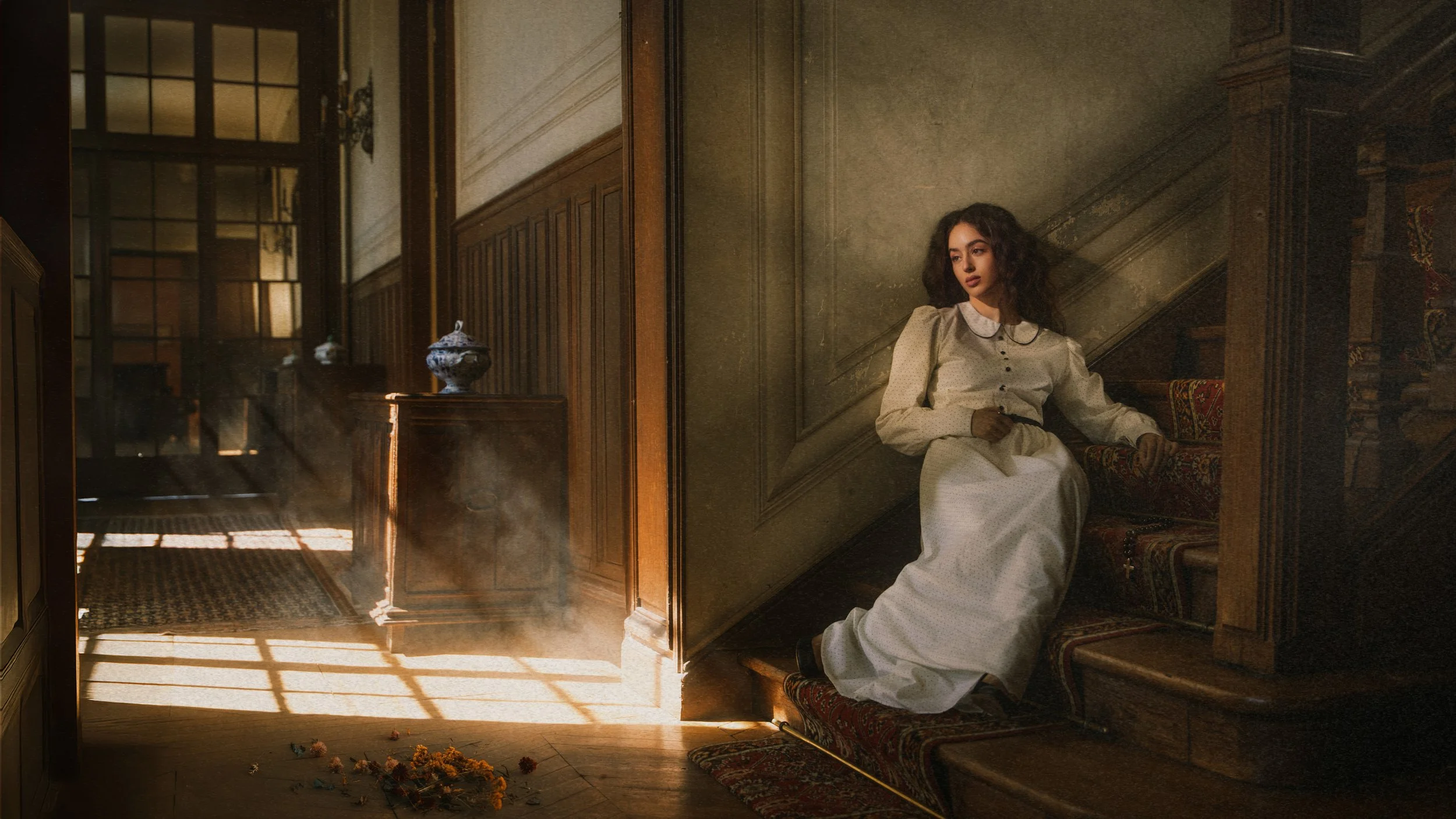 Une femme vêtue d'une robe blanche assise à l'étage d'un escalier en bois dans un intérieur ancien, avec des rayons de lumière passant par une fenêtre et créant des motifs d'ombre sur le sol.
