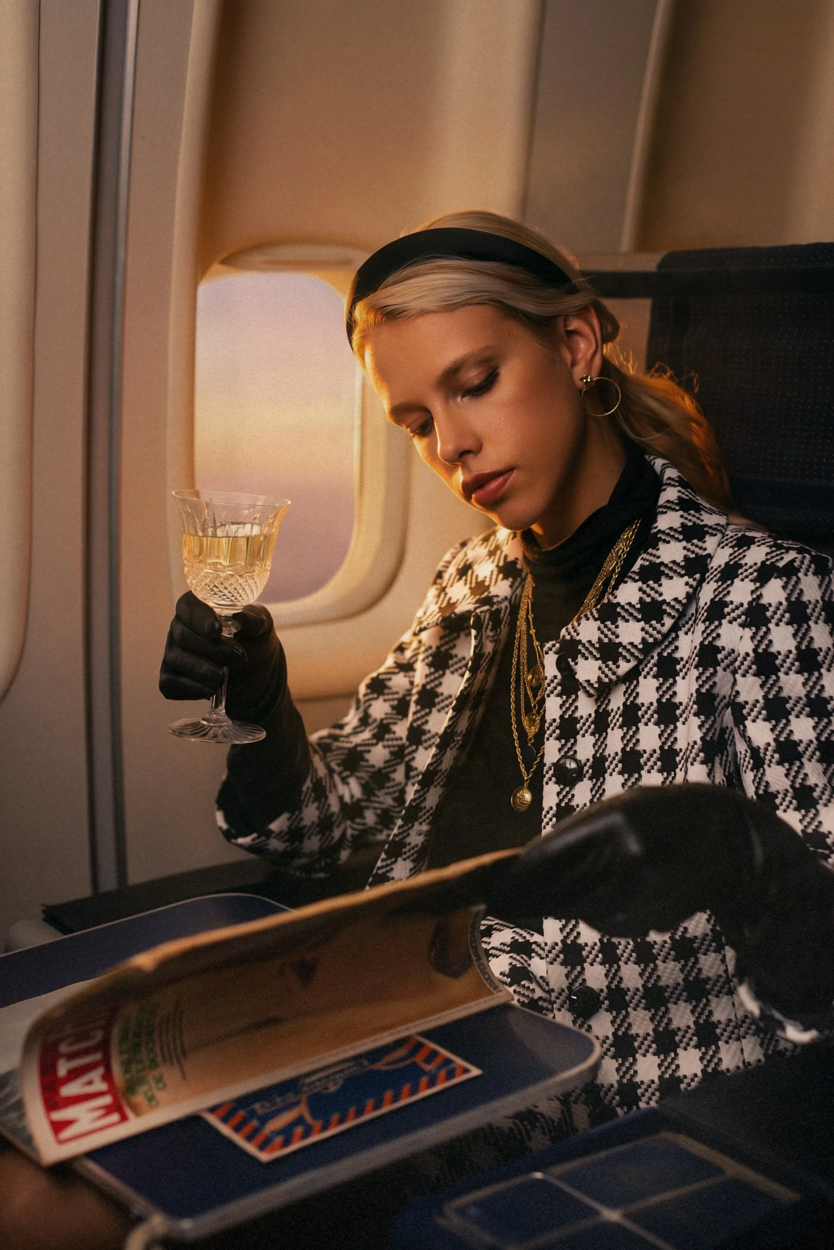 Une jeune femme assise dans un avion, portant des vêtements à motif écossais, une boucle d'oreille en forme de cercle, une chaîne en or, et tenant un verre de champagne. La fenêtre de l'avion laisse passer la lumière du coucher de soleil.