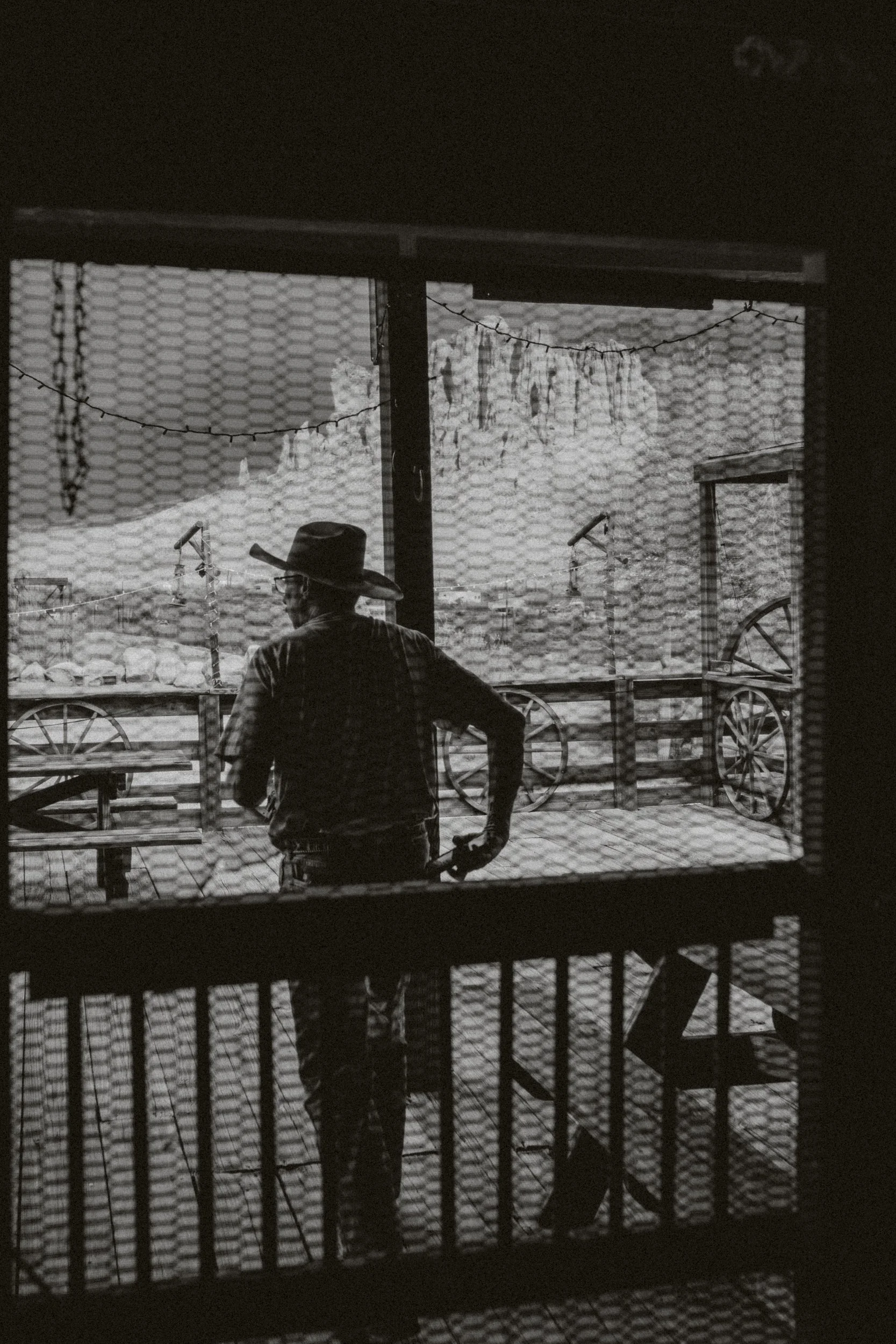 Vue d'une personne portant un chapeau, vue de derrière à travers une fenêtre grillagée, avec des éléments de style vintage comme des roues de chariot et un décor de ferme