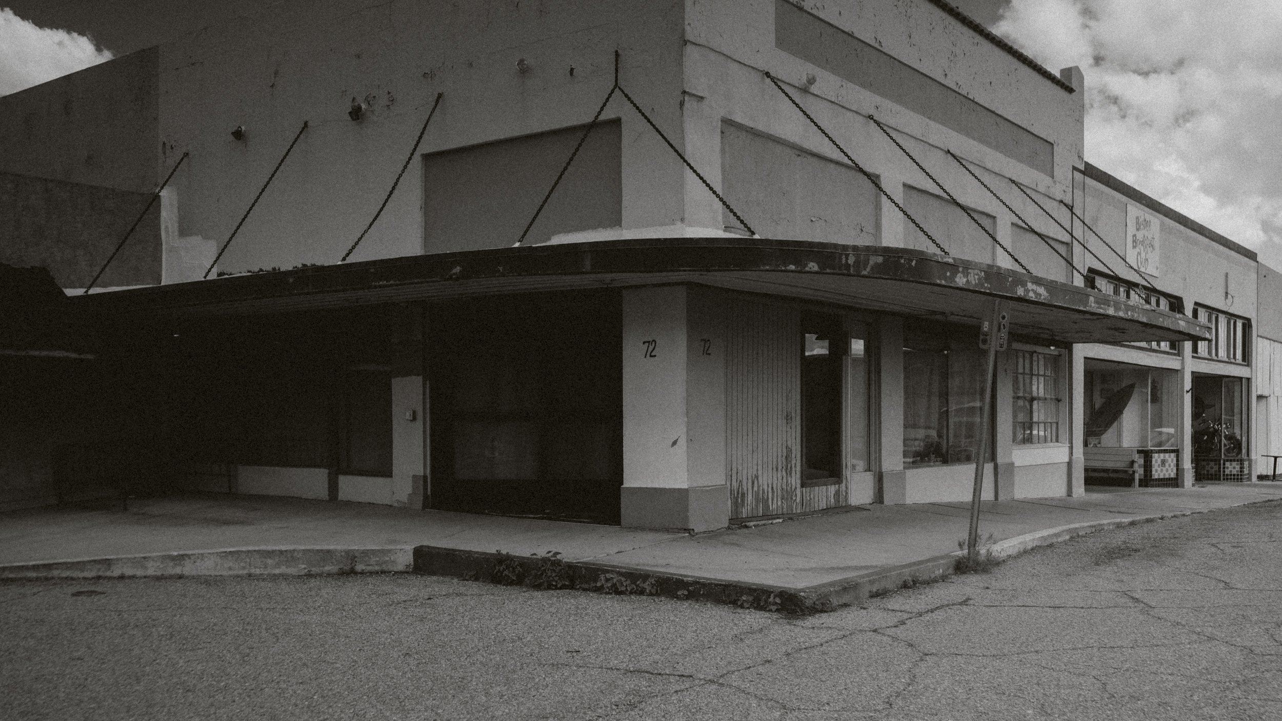 Bâtiment commercial abandonné avec façade en béton blanc, fenêtres cassées, porte rouillée, et un panneau de signalisation abandonné devant.