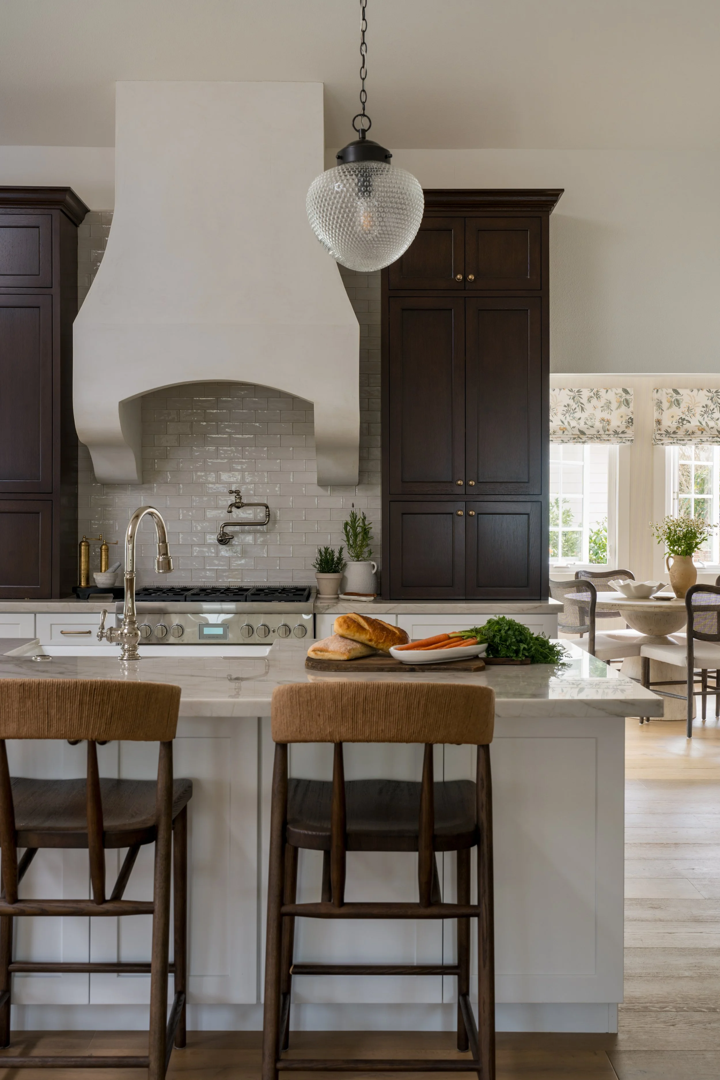 Elegant kitchen with dark wood cabinets, white range hood, globe pendant light, marble island, bar stools, bread, and vegetables