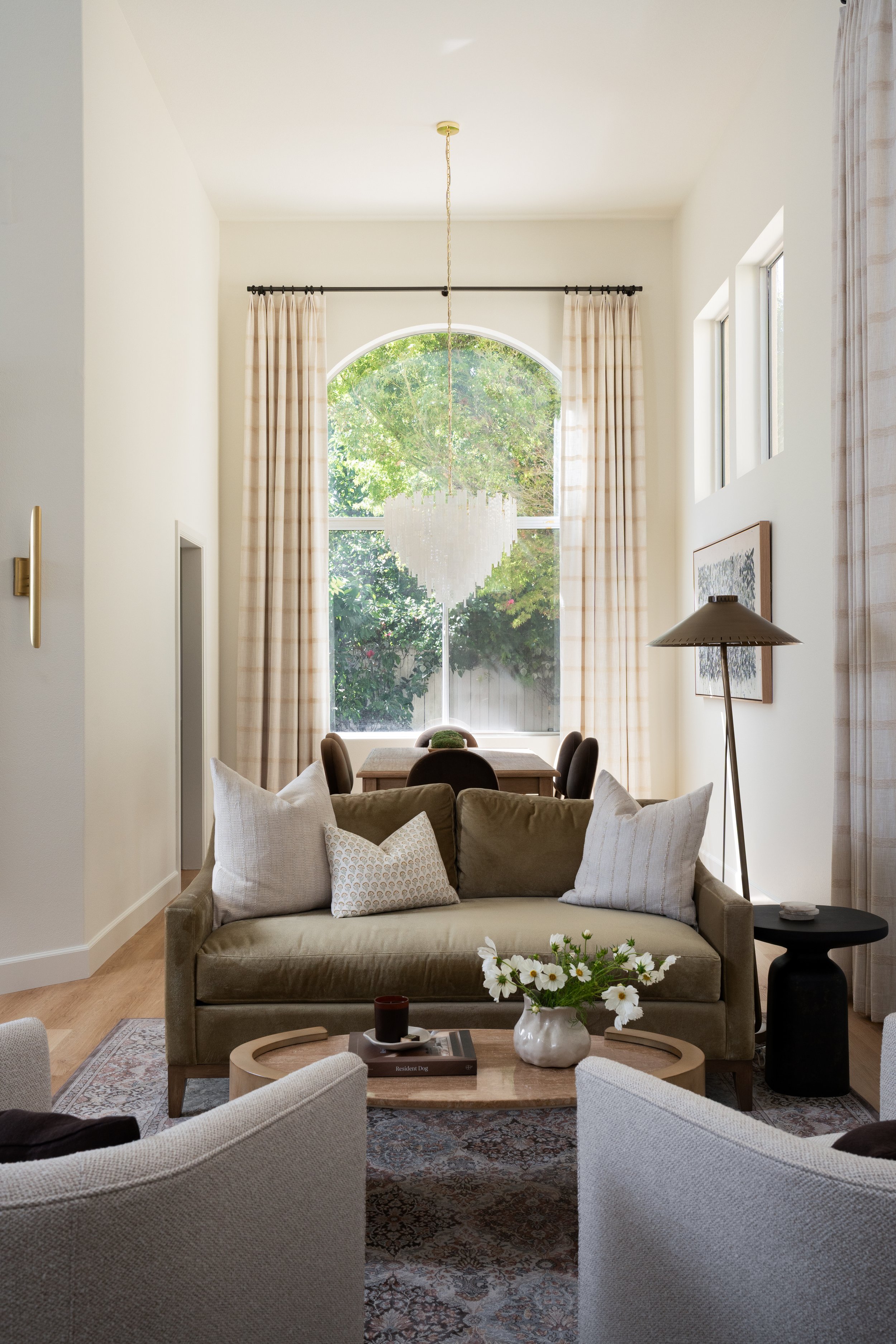 Bright modern living room with olive sofa, patterned pillows, arched window, dining table, pendant light, floor lamp, and floral coffee-table arrangement