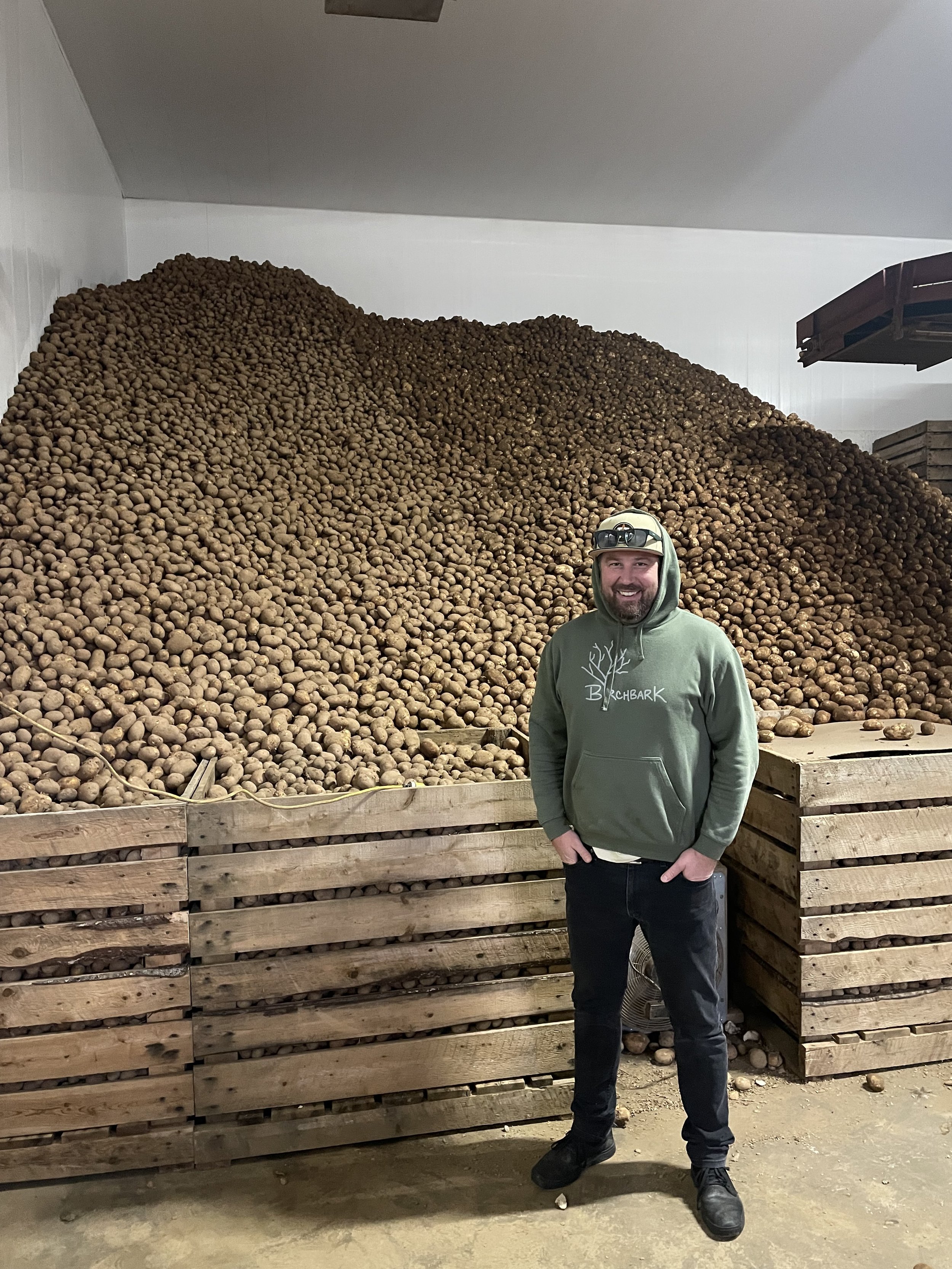 A man in a green hoodie with a ball cap and sunglasses poses smiling in front of, literally, enough potatoes to feed an army.