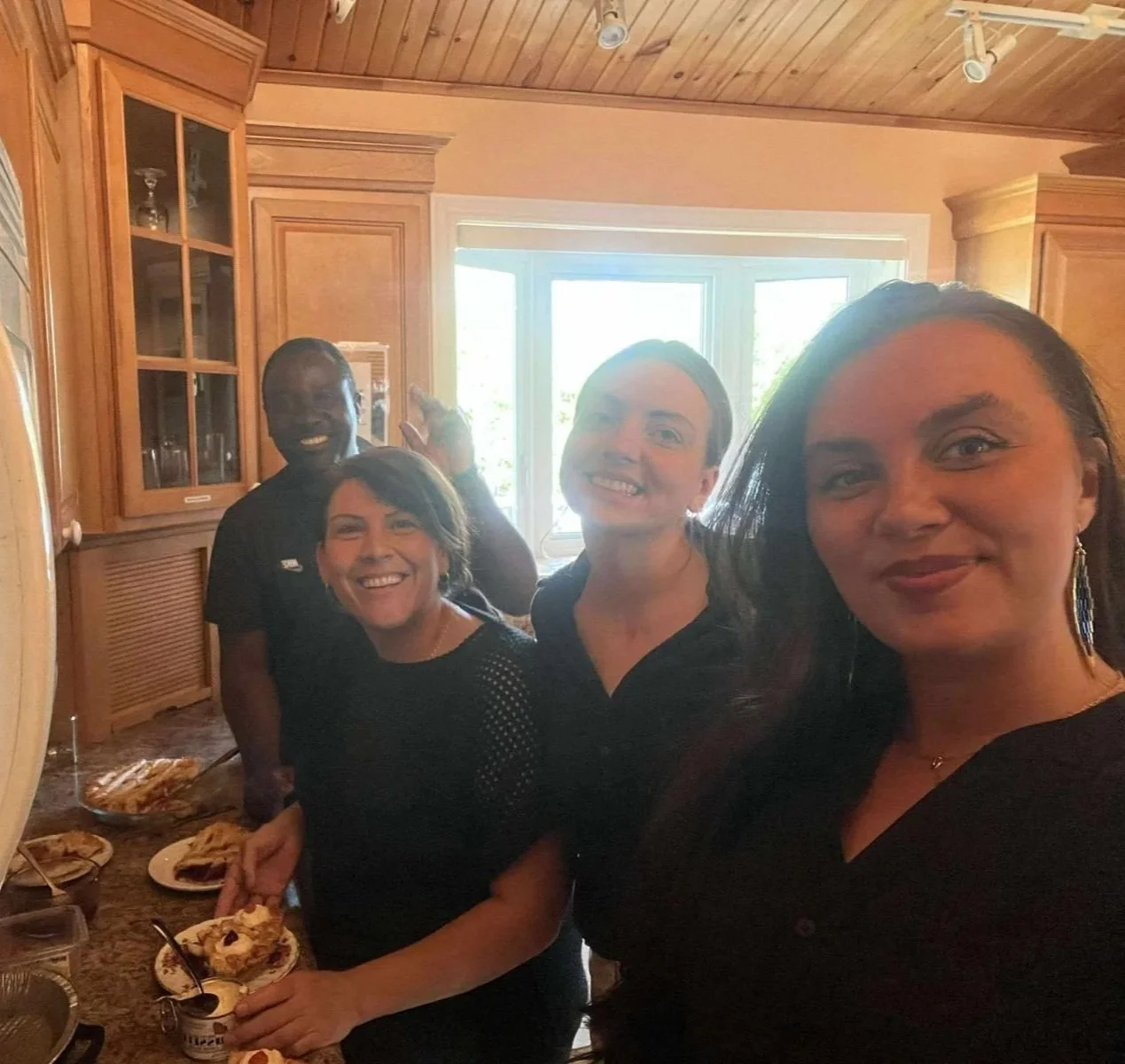 Four smiling people in a kitchen serve up plates of pie.
