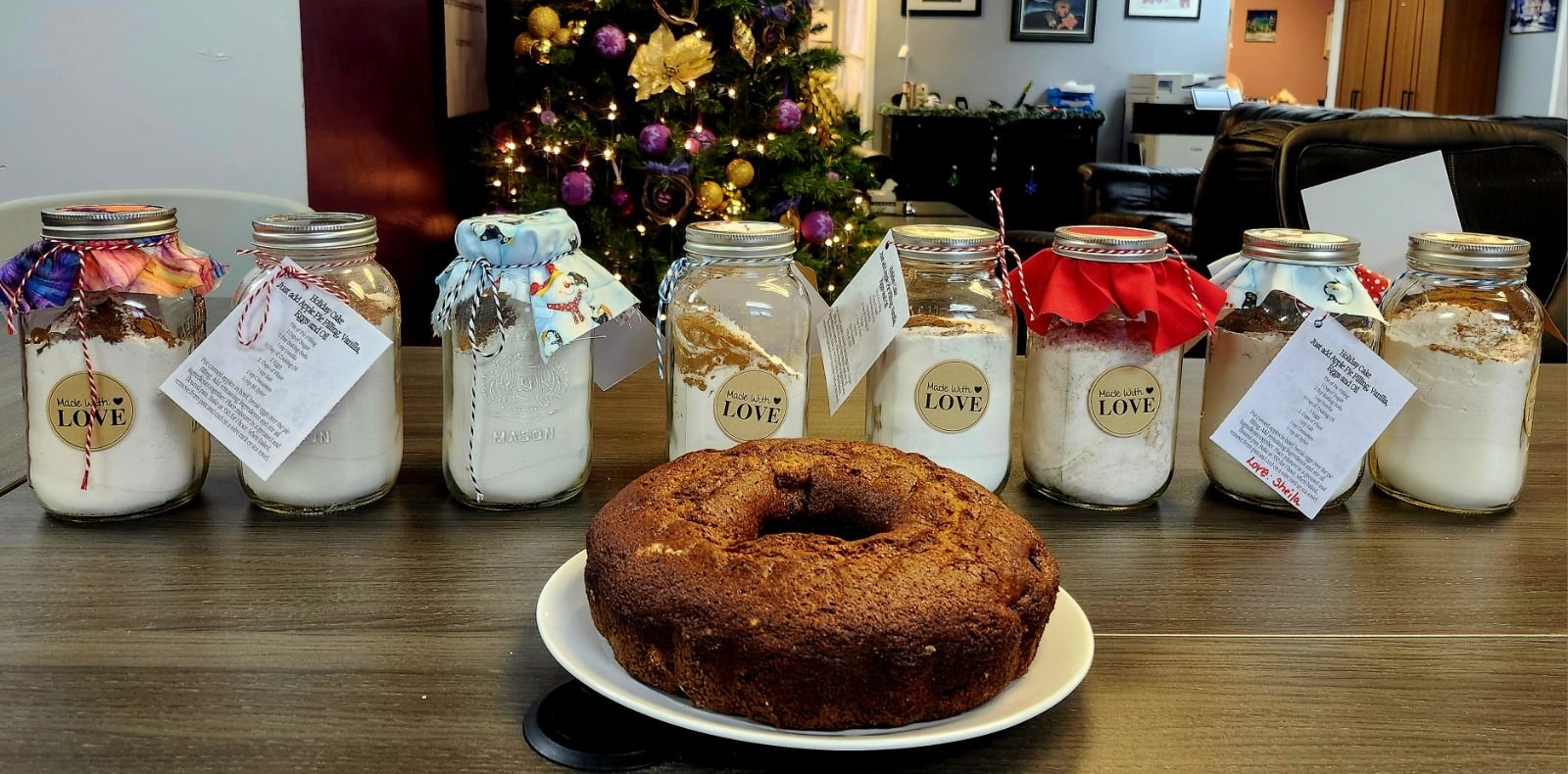 A freshly-baked bundt cake in the foreground of a cozy Christmas room, with decorated jars of cake mix lined up behind the cake.