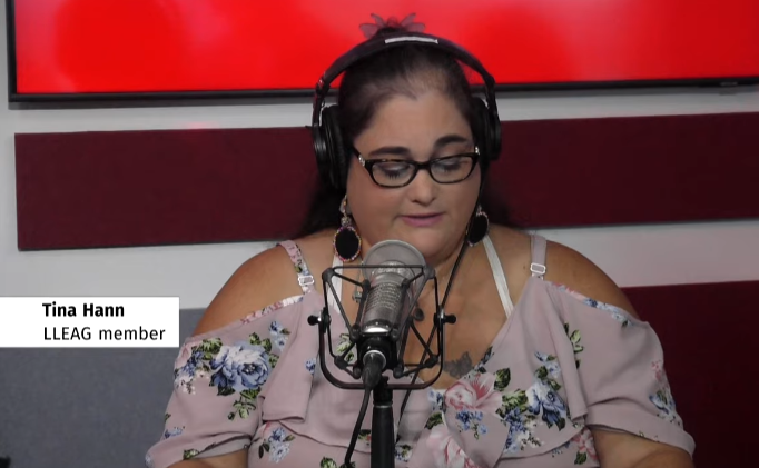 A woman in a cold-shoulder blouse, glasses, and dangly earrings speaks in a radio studio.