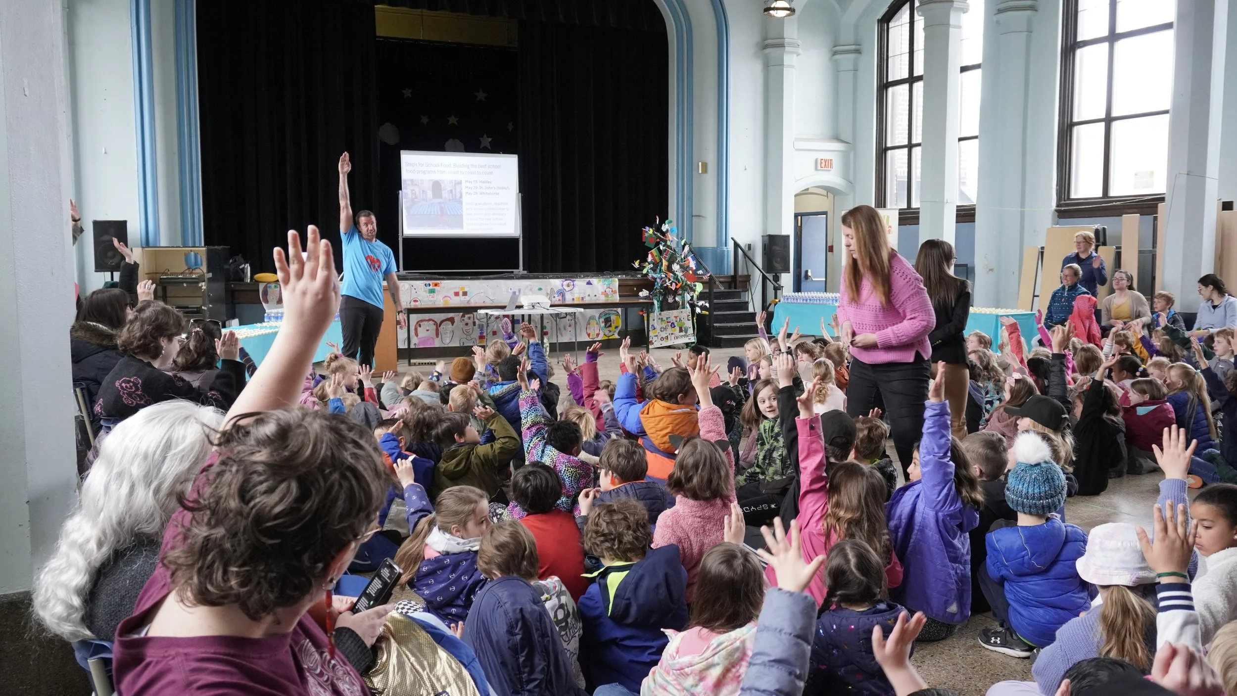 An auditorium full of kids and teachers listening to a man in a blue shirt speaking with his hand up at the front of the room.