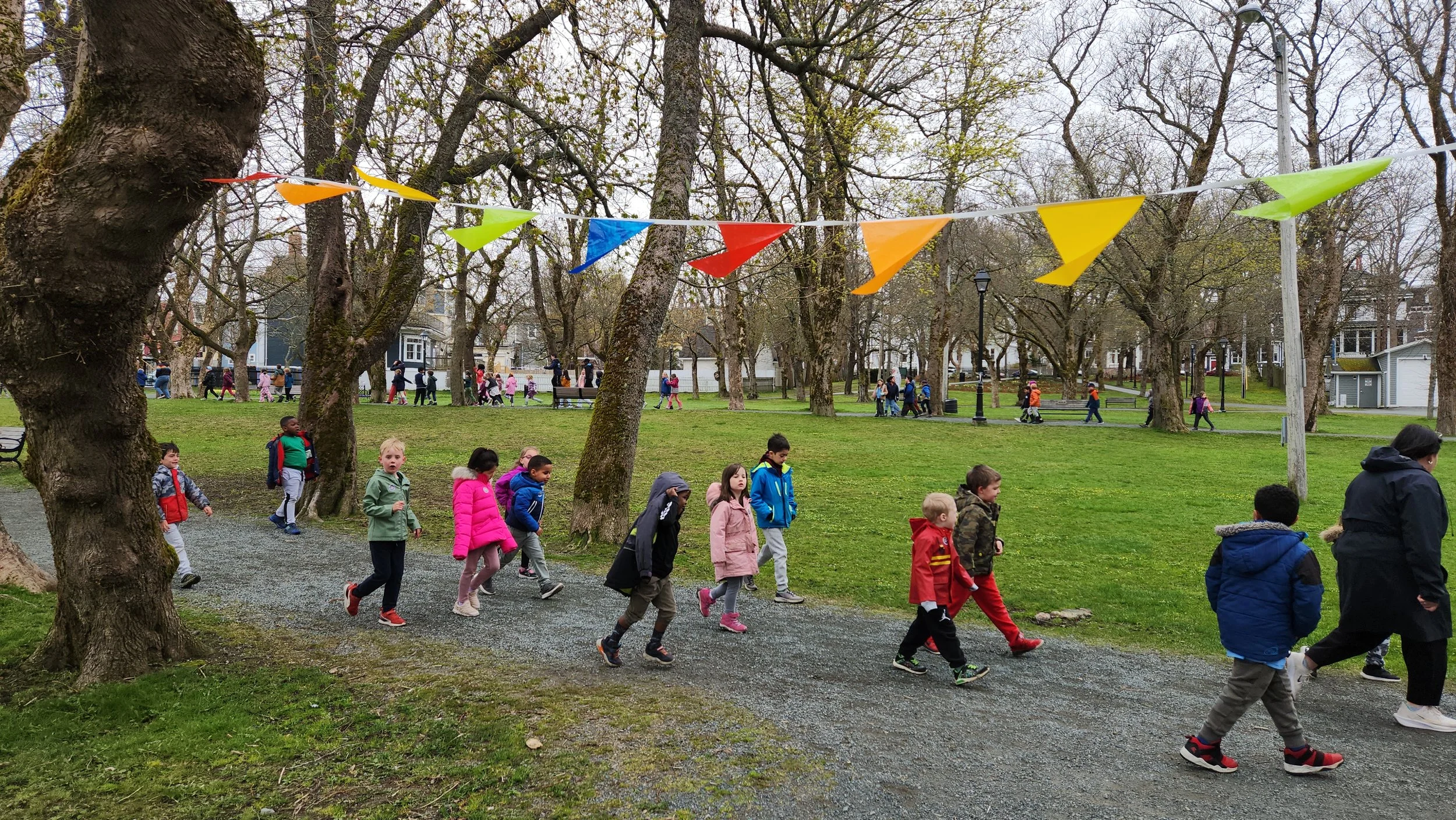 Small groups of children walk together through Bannerman park amidst colourful hanging buntings on a cloudy day.