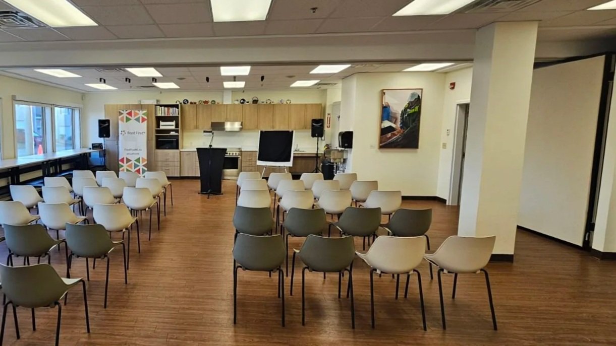A view of the main room at Our Table post-renovation: the back wall has been transformed into a kitchen, art is on the walls, and a fleet of chairs are arranged for a presentation with a podium set up.