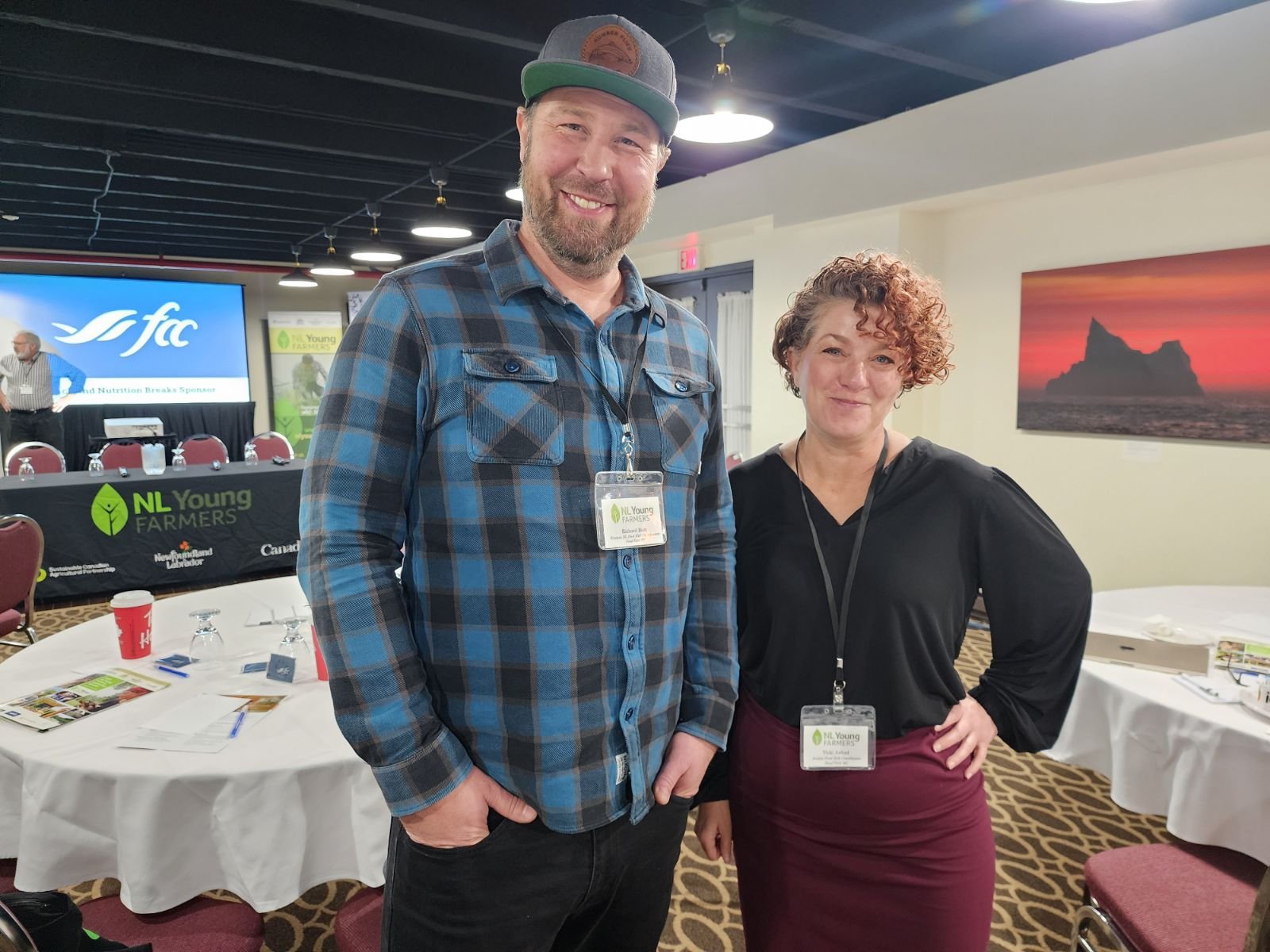 A fall man in flannel and a ball cap poses with a shorter woman in a black blouse in a conference room. Both are smiling and wearing lanyards.