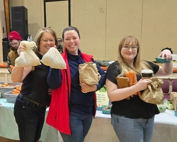Three Food on the Move customers hold up their haul of produce in cloth and paper bags, smiling.