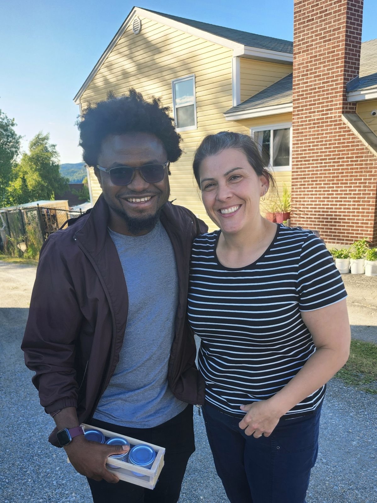 Rasheed, a Ghanaian man in sunglasses and Dana, an Indigenous woman in a striped shirt, pose together smiling outside a residence.