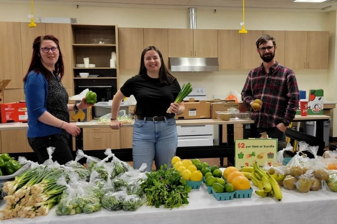 Three Food First NL staff pose with produce behind a Food on the Move spread at Our Table/Charter Avenue.