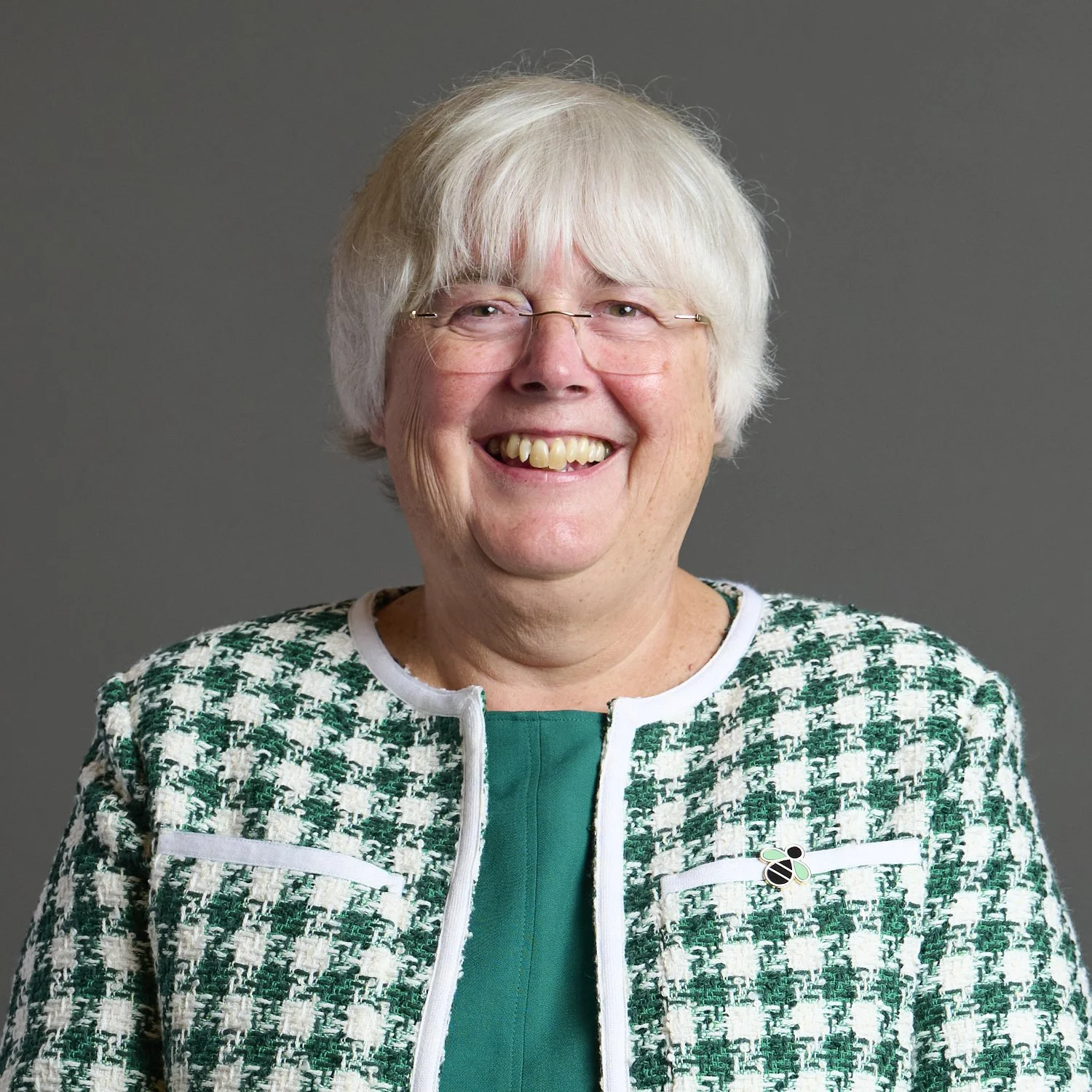 The official portrait of Charlotte Cane - a head and shoulders photo: she wears a green and white checkered cardigan, has a white bob haircut and glasses and is smiling.