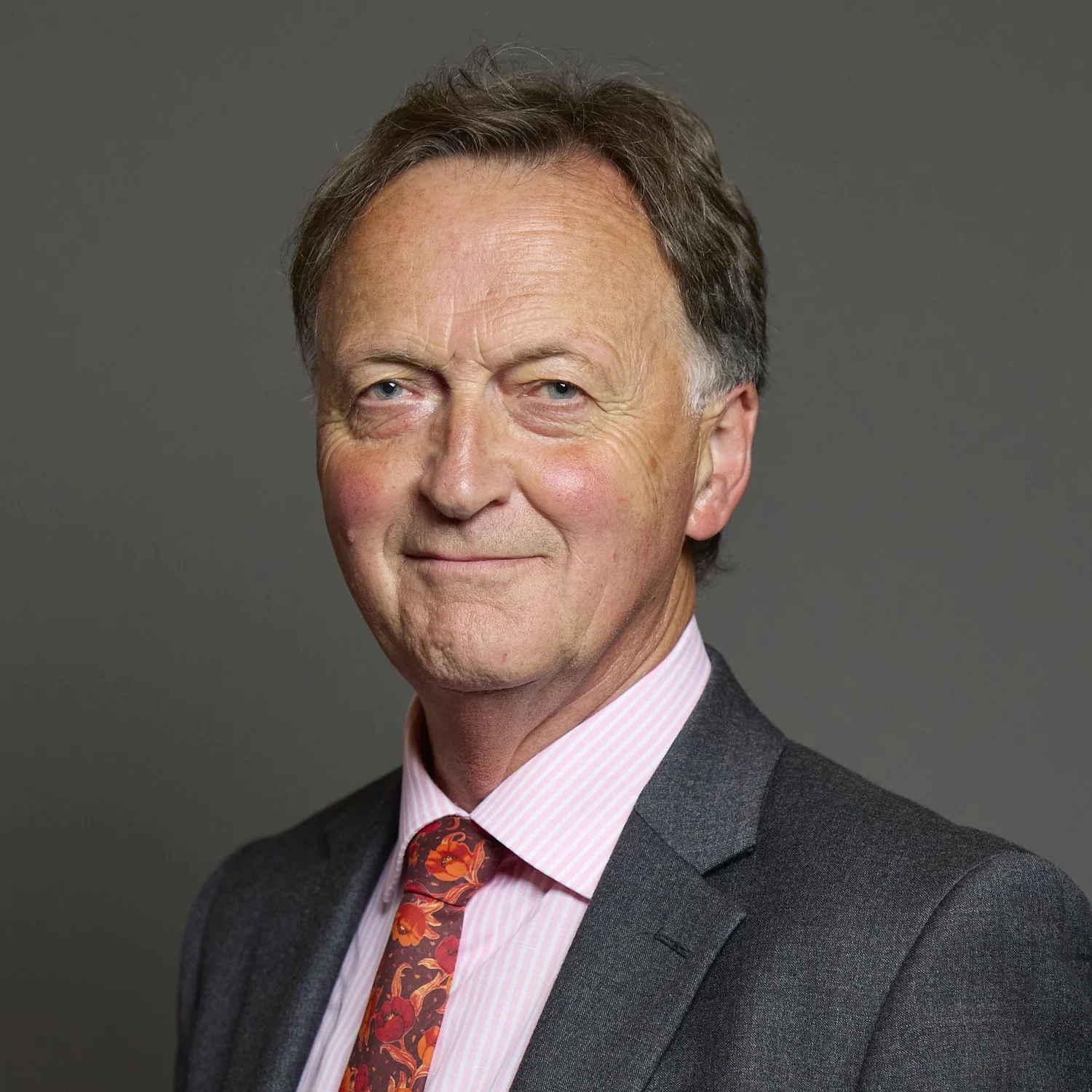 Portrait of Andrew George MP.  A head and shoulders framing, he stands slightly at an angle. He is smiling, has a grey suit jacket, pink shirt, and red floral tie.