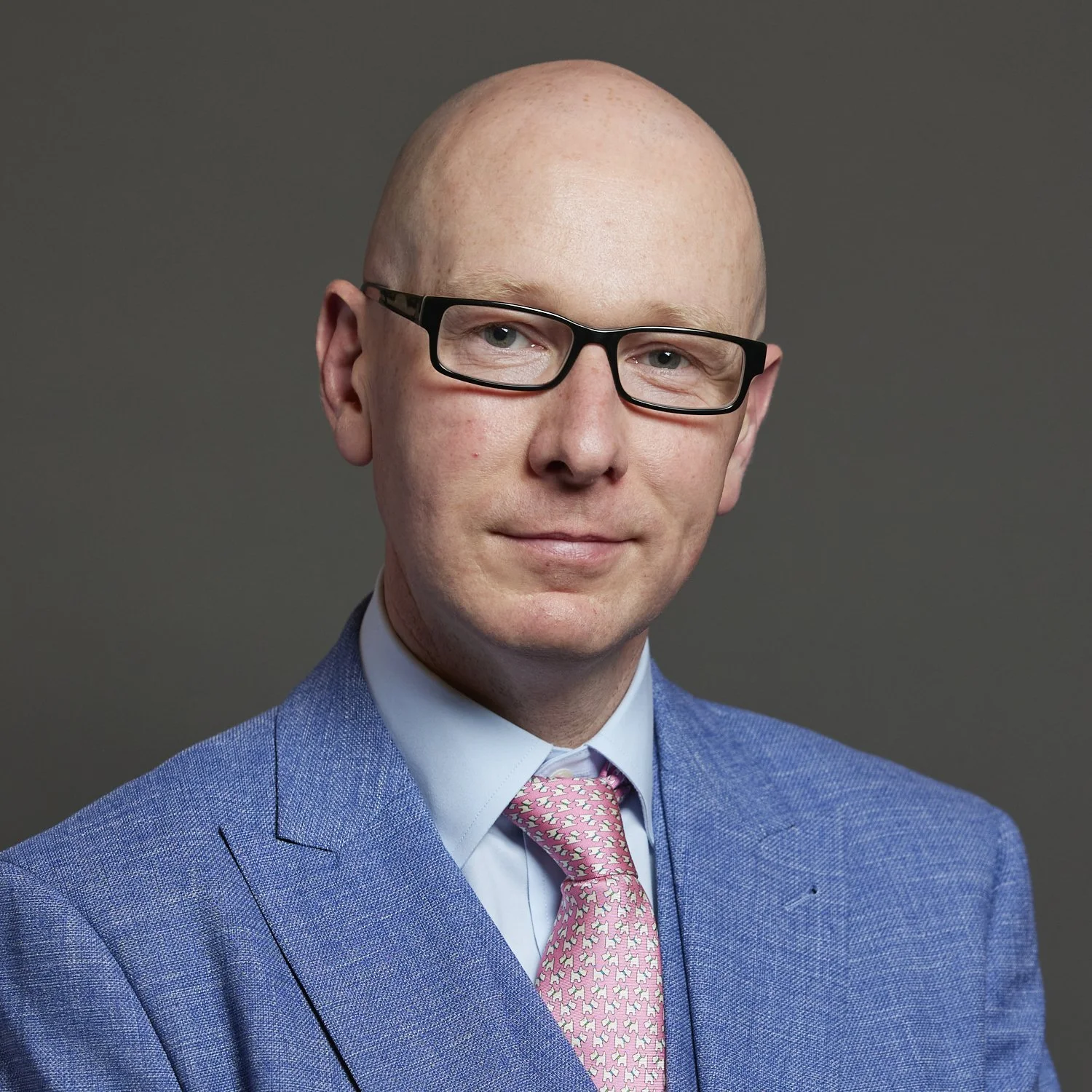 Official portrait of Patrick Hurley MP. A head and shoulders photo - he wears a blue suit jacket and pink tie, he is bald and has small thick rimmed glasses.