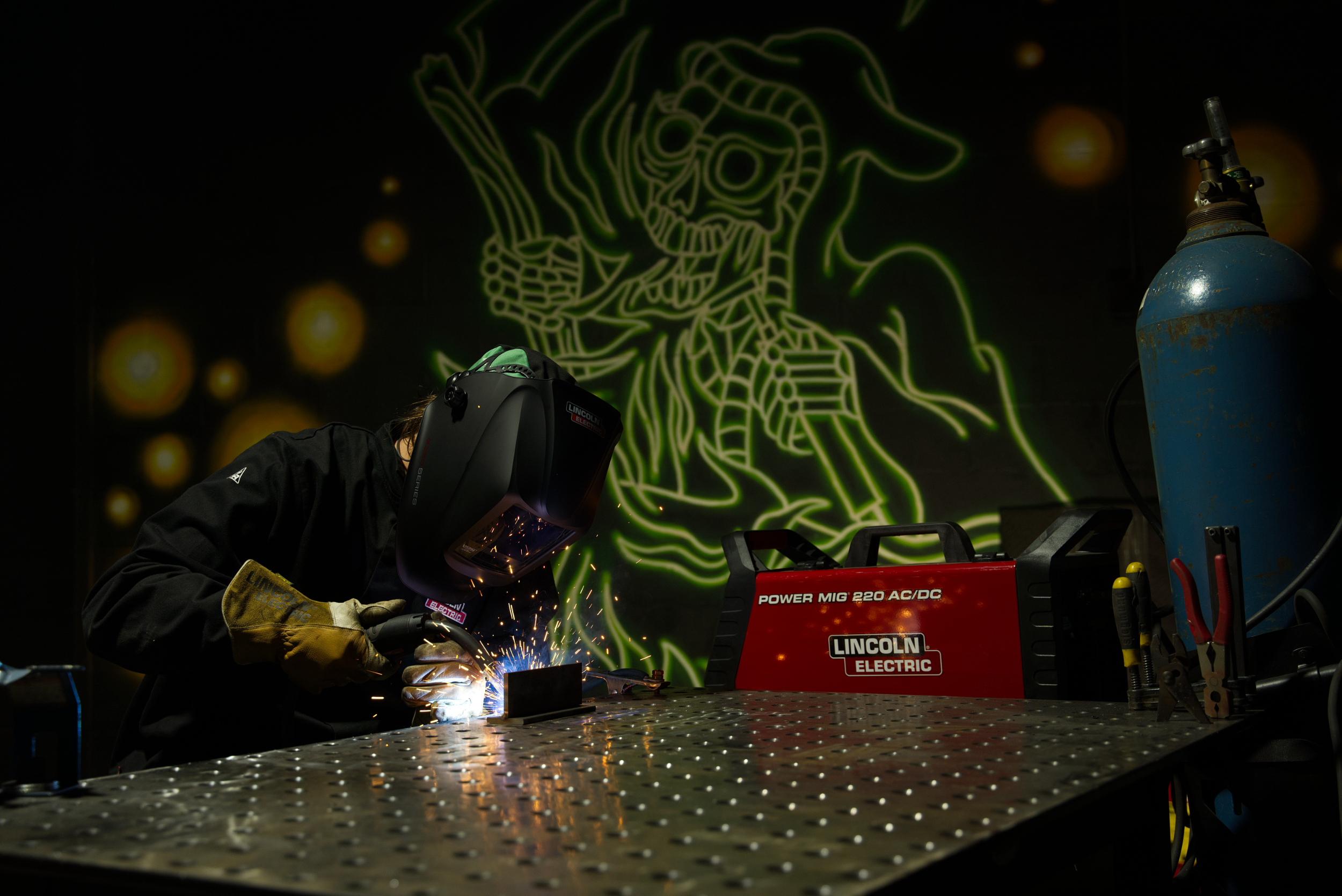 Person welding in a workshop, wearing protective gear and helmet, with bright sparks and blue exhaust ventilation hoses.