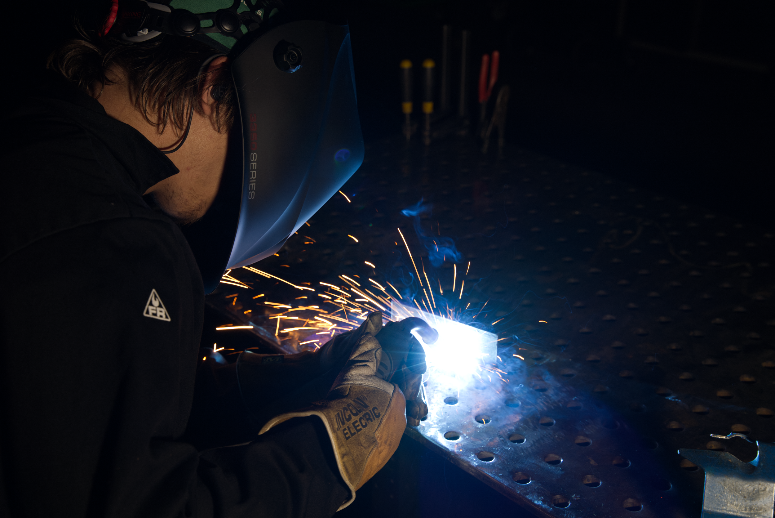 Person wearing a welding helmet and protective clothing, working at a welding station with tools, sitting on a stool. Background features colorful graffiti and a fire extinguisher is visible.