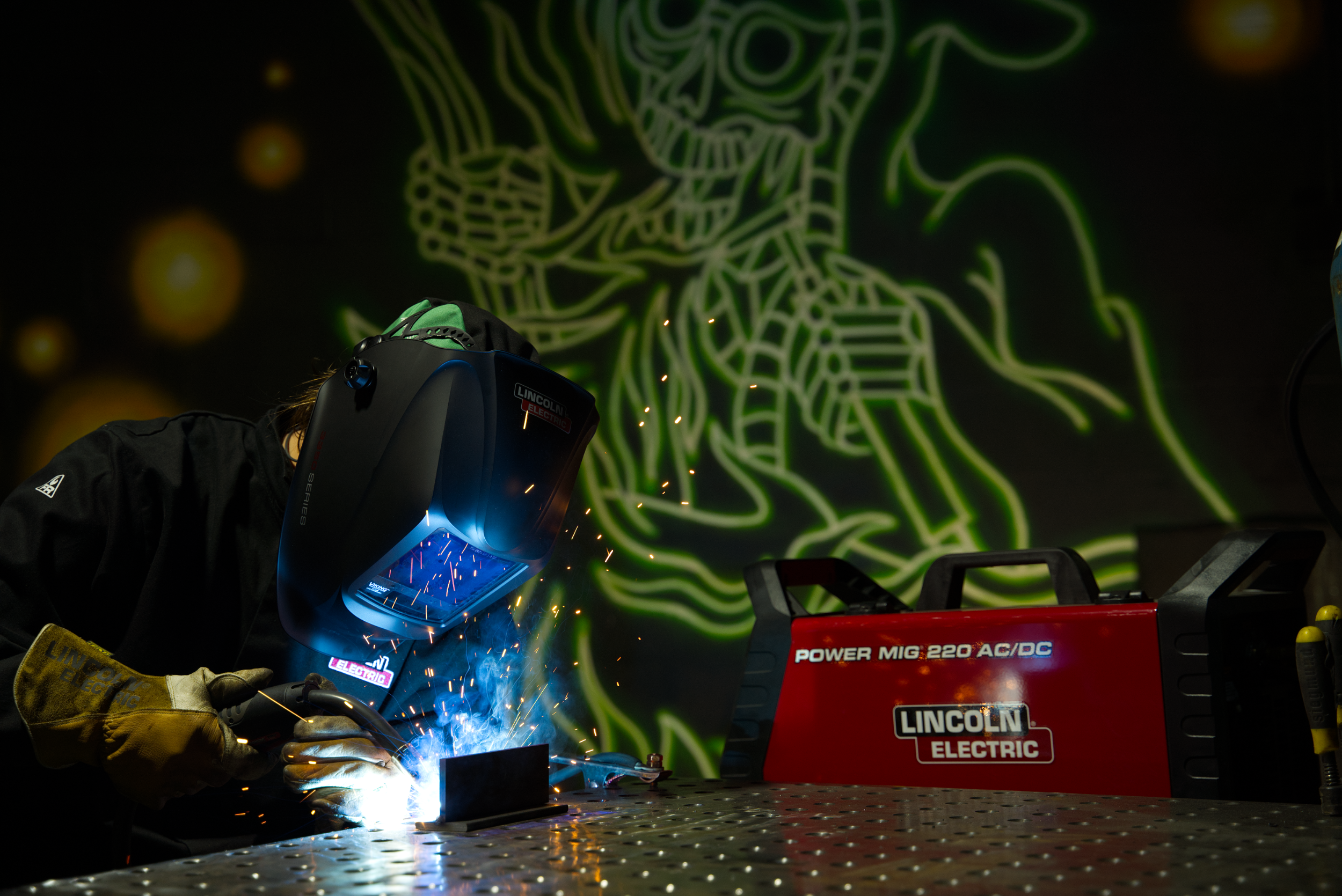 Welder wearing protective gear using a welding torch, with bright sparks and colorful graffiti in the background.