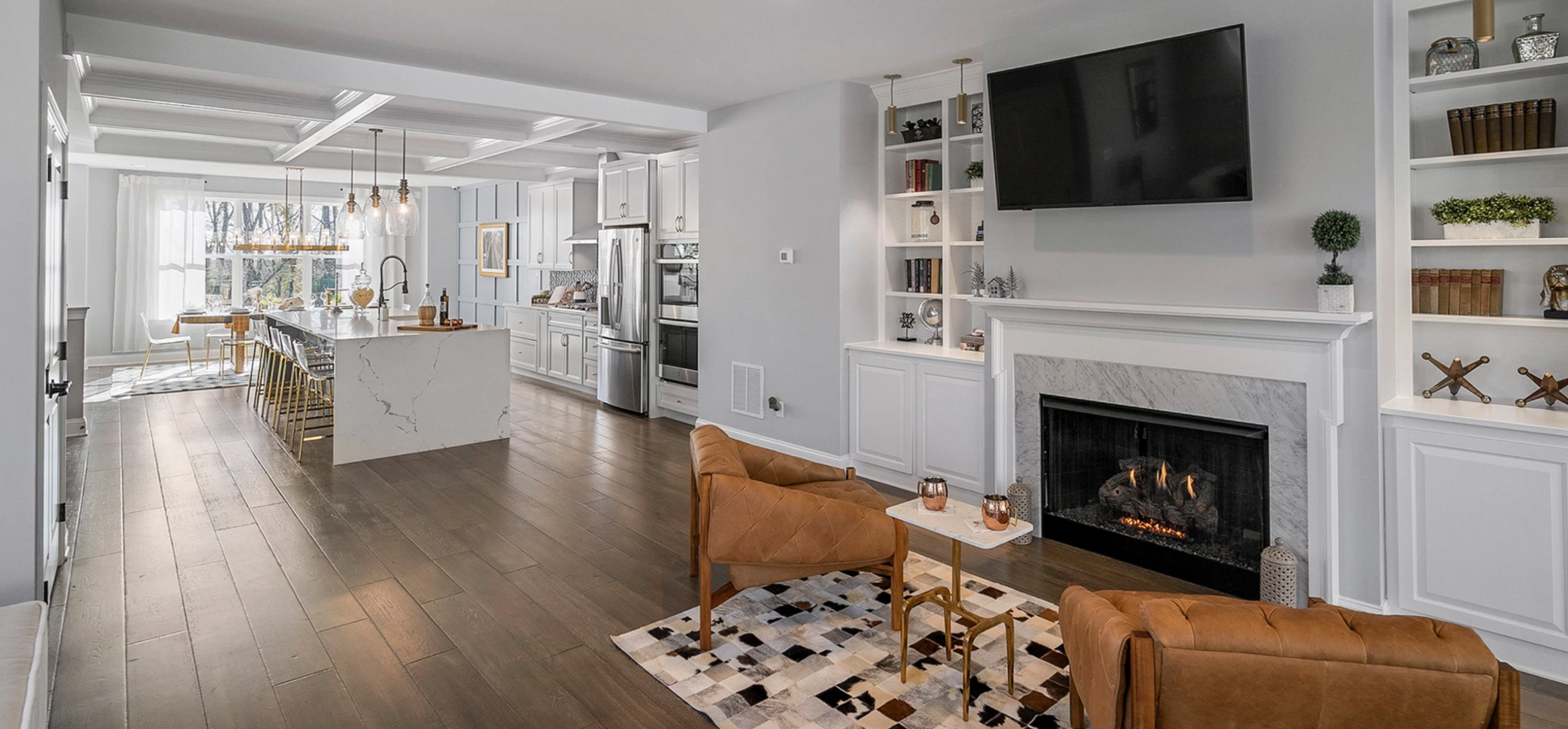 Open-concept living room and kitchen with white cabinetry, stainless steel appliances, wooden flooring, a fireplace with a mounted TV above, built-in shelves, a seating area with two tan leather chairs, a small coffee table, and decorative items.