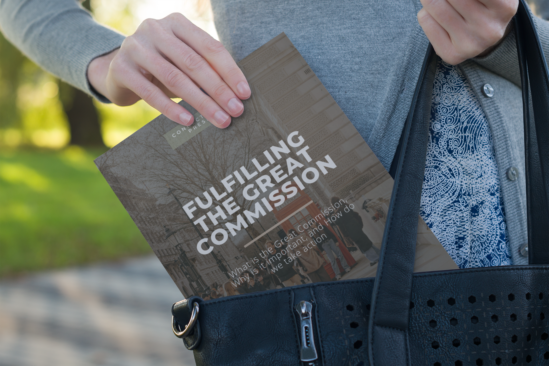 Person wearing a gray sweater and patterned blue shirt, placing a flyer about city planning into a black handbag outside on a park bench.