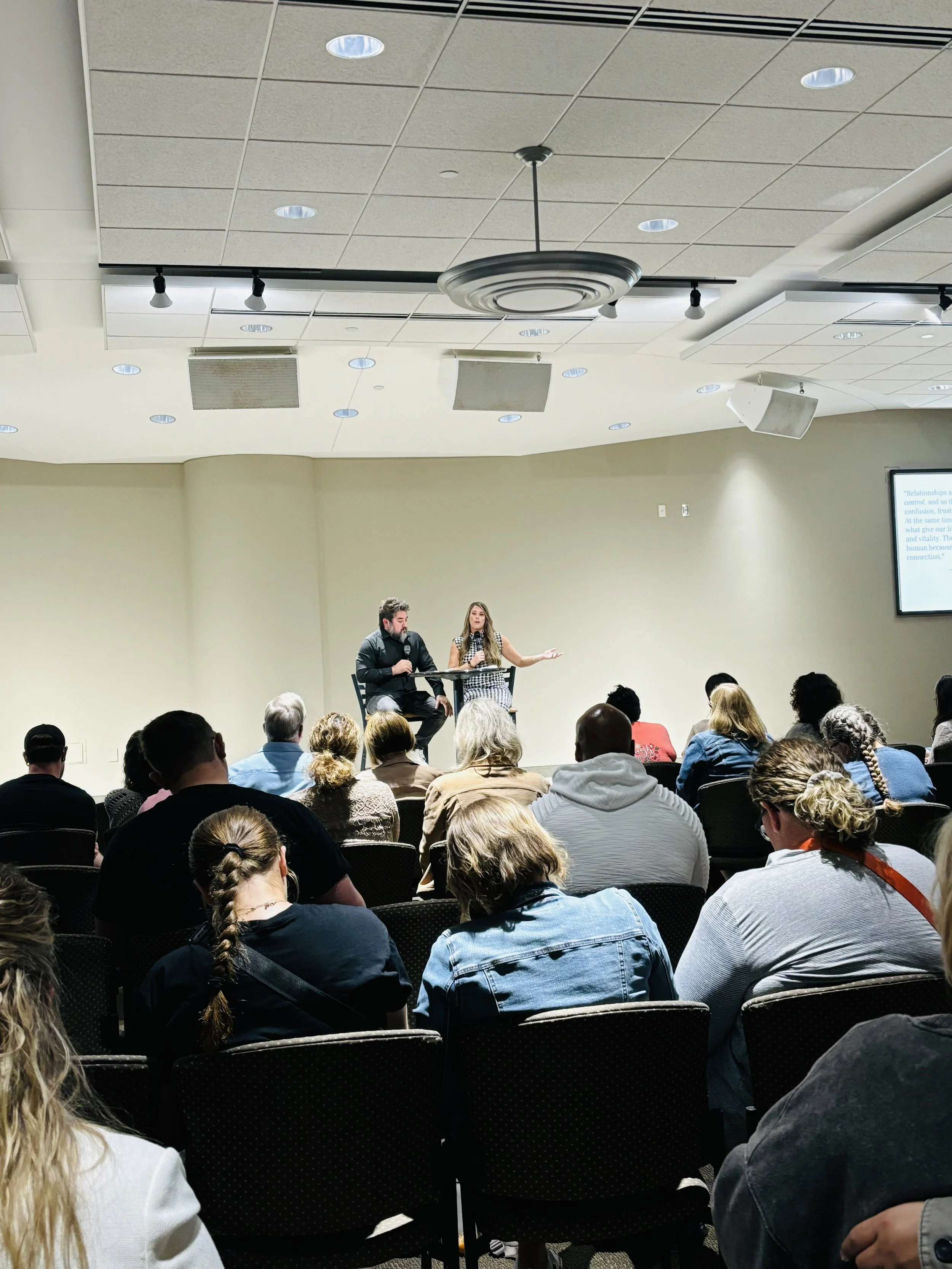 Javier and Danielle Mendoza, speaking on stage during a presentation in front of an audience, with a screen displaying text to the right.
