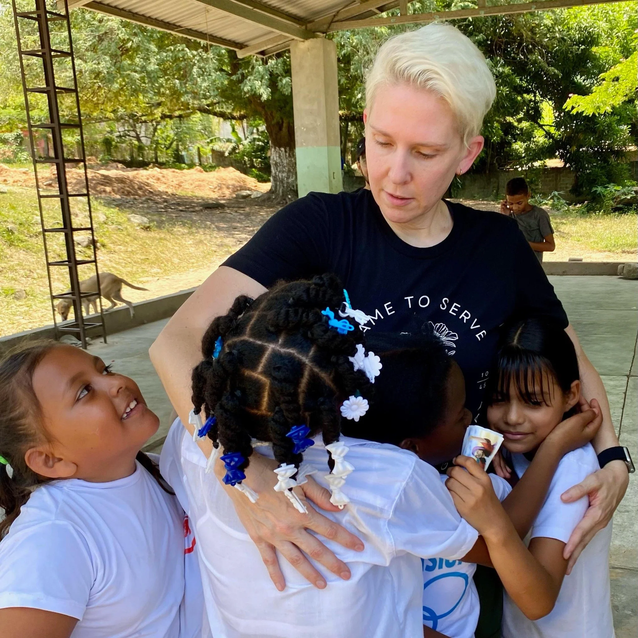 A woman with short, platinum blonde hair hugging and comforting four young girls outdoors under a roofed area, with trees and a boy in the background.