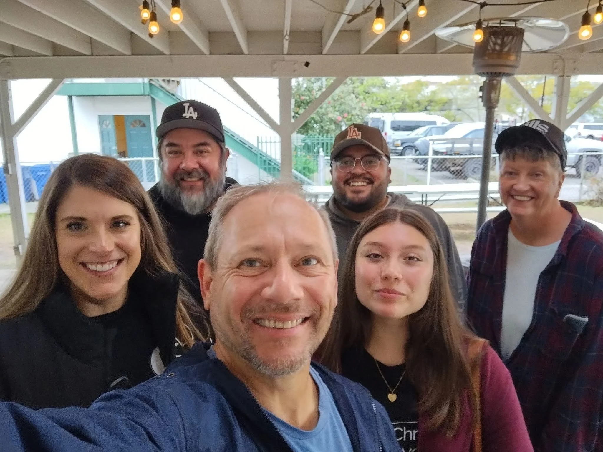 Group of seven smiling people taking a selfie under a covered outdoor area, with string lights hanging above and parked cars in the background.