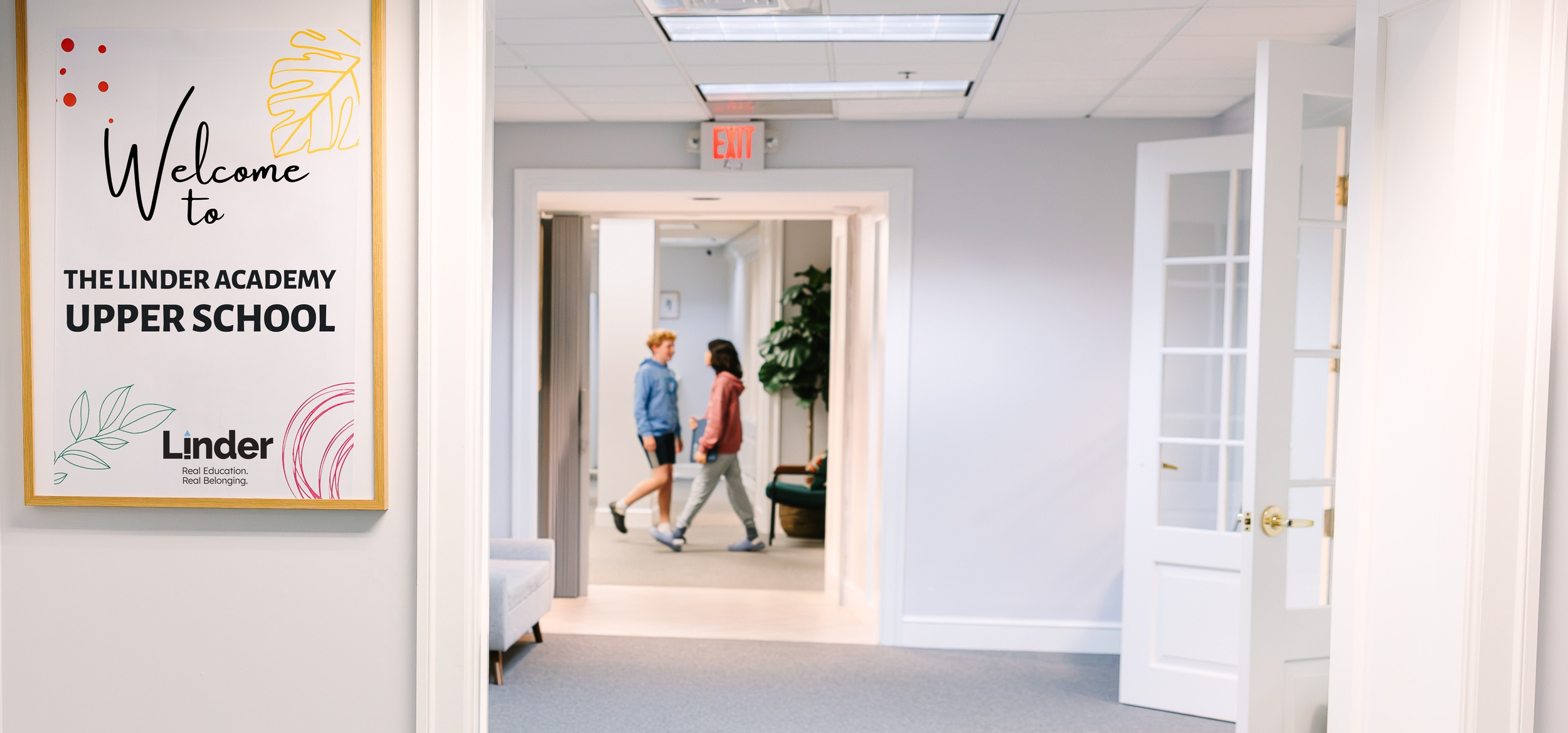 Interior of a school hallway with a welcome sign for The Linder Academy Upper School on the left and two students walking and talking in the background.