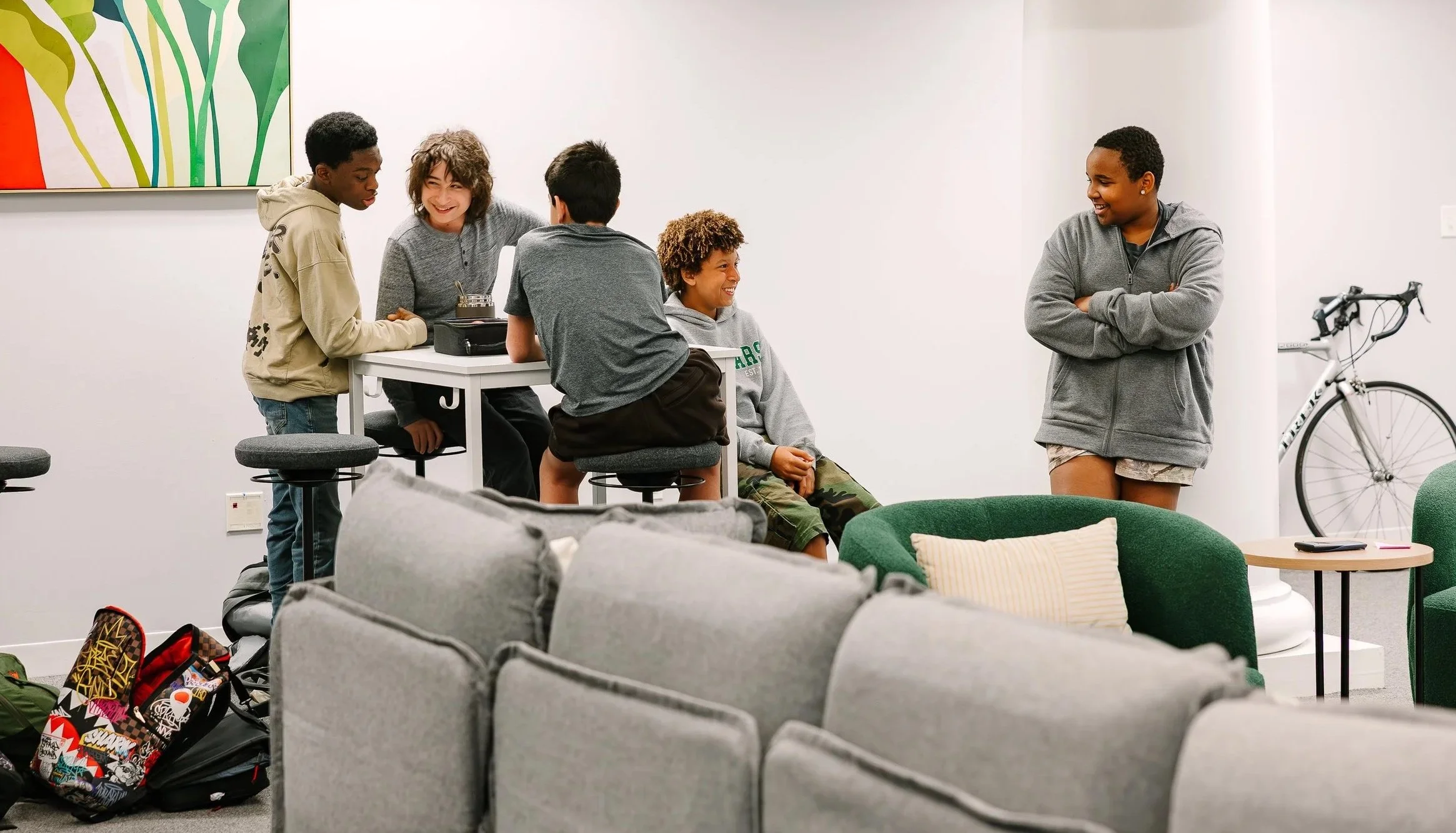 Group of five teenagers in a casual lounge area, some sitting on stools and others standing, smiling and talking. Three of them are gathered around a high table, while one stands with arms crossed smiling at the others. In the background, a bicycle leans against a wall, and in the foreground, there are gray couches and a green chair with a pillow.