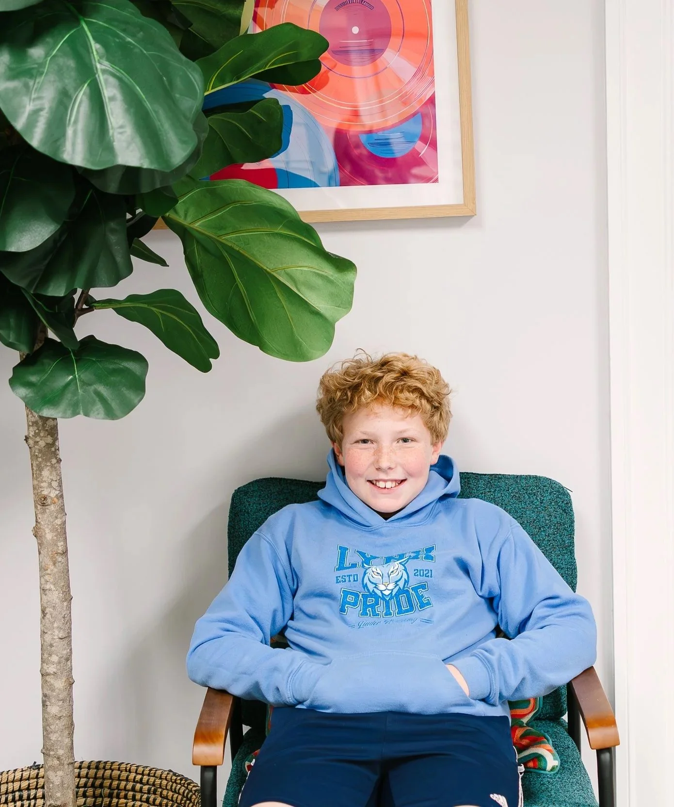 A smiling boy with curly red hair wearing a blue sweatshirt sitting in a green armchair next to a large indoor plant and colorful artwork on the wall.