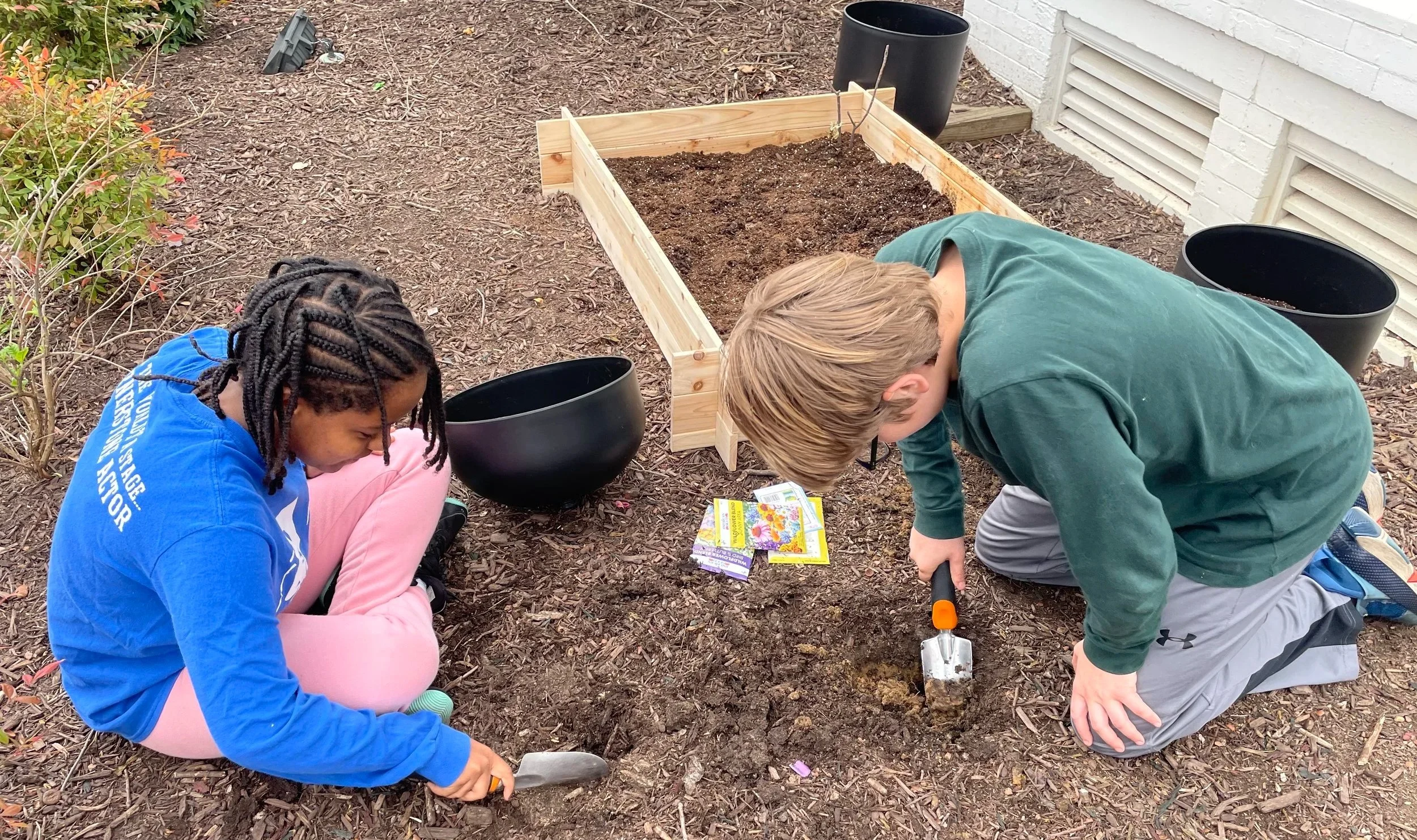 Two children planting in a garden bed. One with braided hair in a blue shirt and pink pants, the other with blonde hair in a green shirt and gray pants, both kneeling and working with small gardening tools.