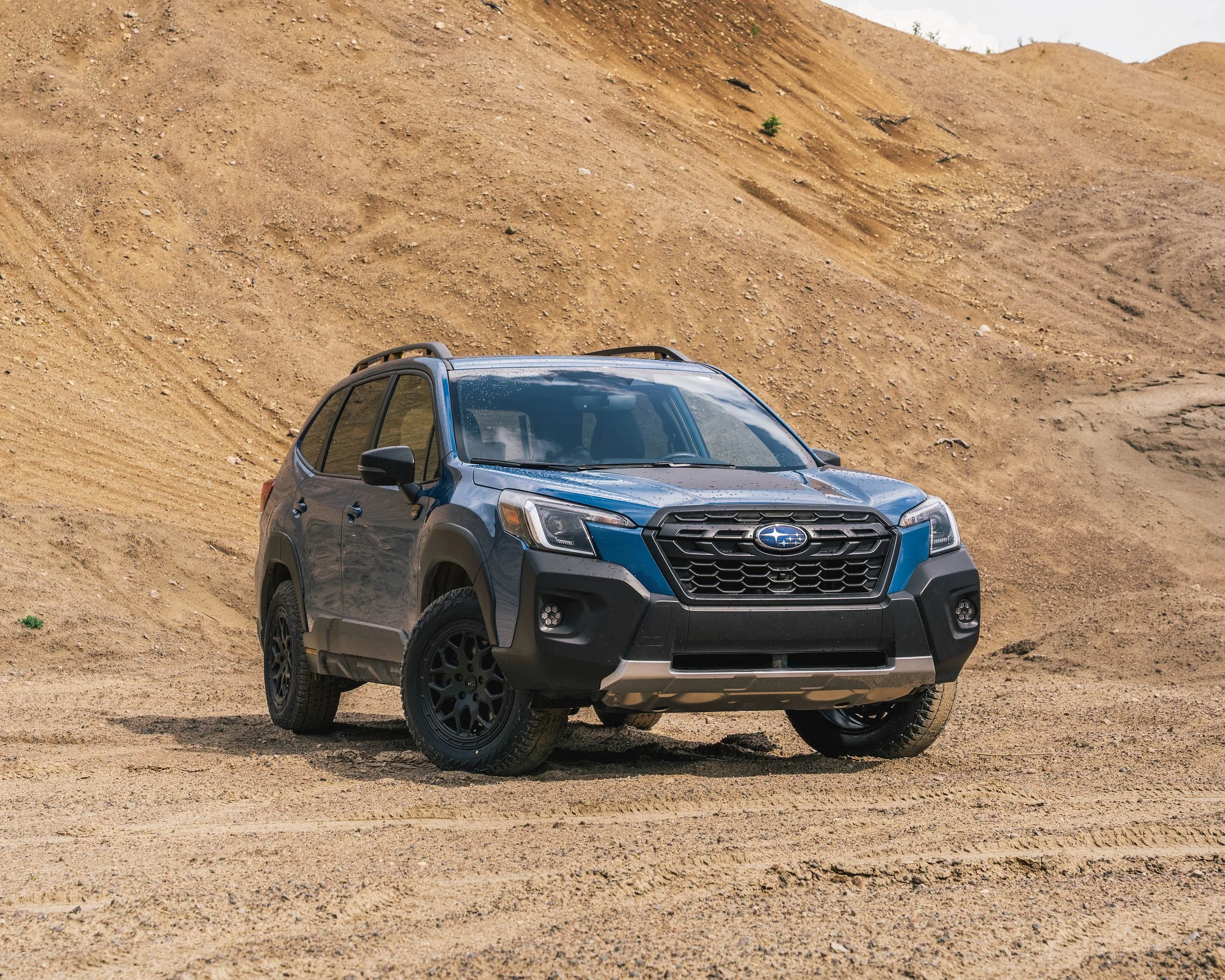 Blue Subaru SUV parked on a dirt terrain with a large sandy hill in the background.