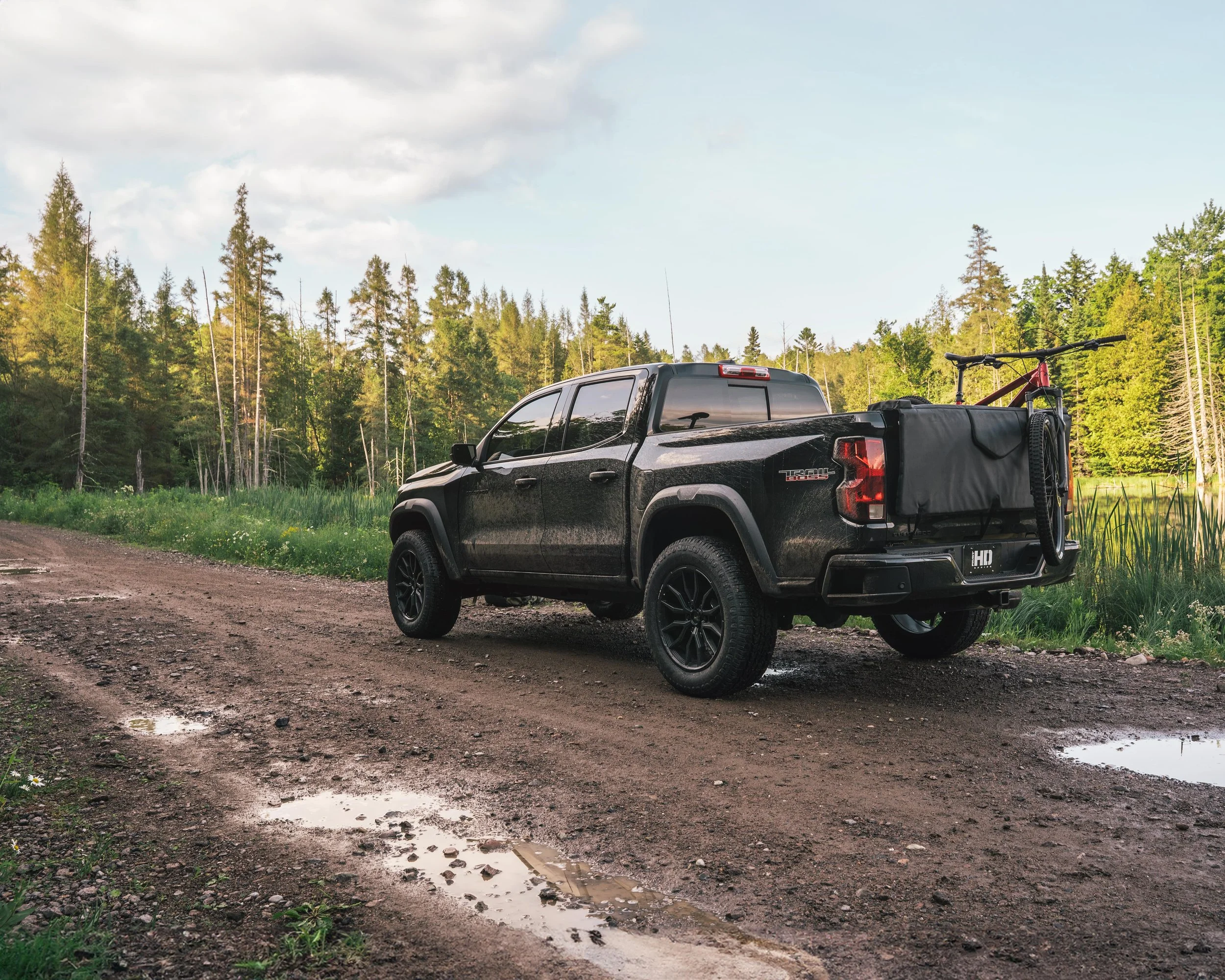 A black pickup truck parked on a muddy dirt road in a forested area with a mountain bike in the truck's bed.