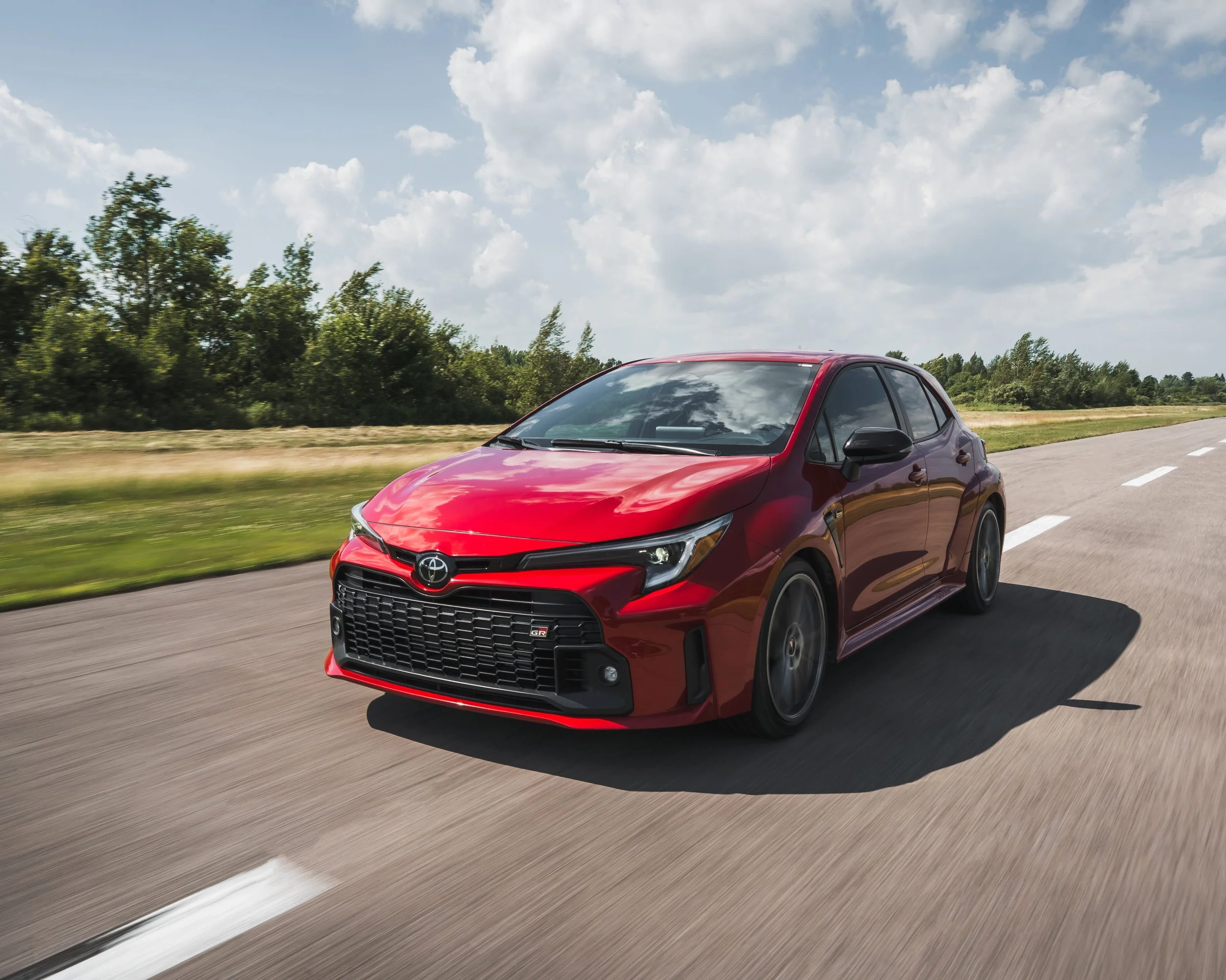 A red Toyota Corolla driving on a highway with a green landscape and blue sky with clouds in the background.