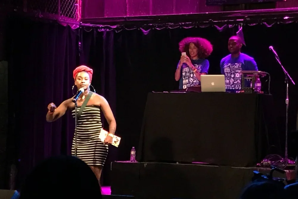A woman with a pink headwrap and striped dress speaking into a microphone on stage, with two people behind a DJ booth and purple lighting in the background.