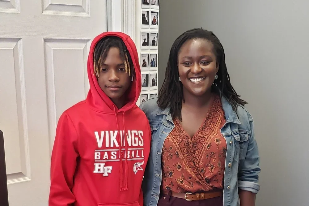 A woman and a teenage boy standing together indoors. The woman is smiling, wearing a denim jacket over a patterned blouse. The boy has a serious expression, wearing a red hoodie with "Vikings Basketball" written on it, and has dreadlocks.