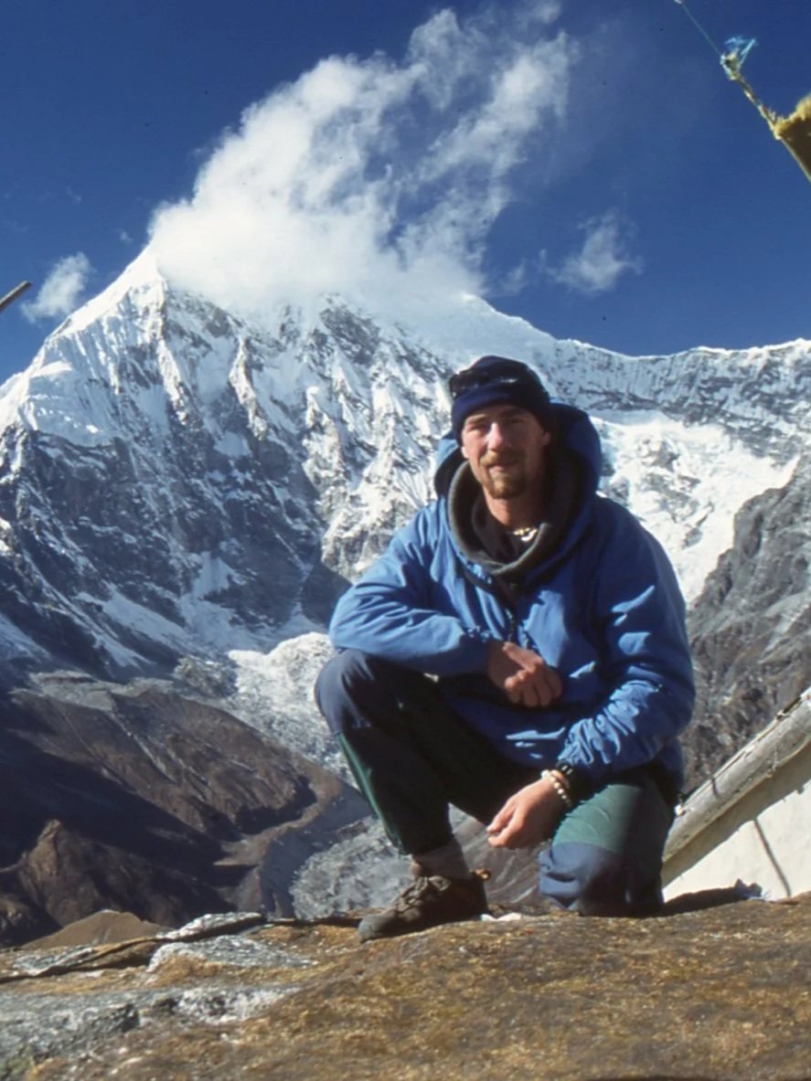 A man in outdoor gear crouching on rocky terrain with snow-covered mountain and glacier in the background under blue sky.