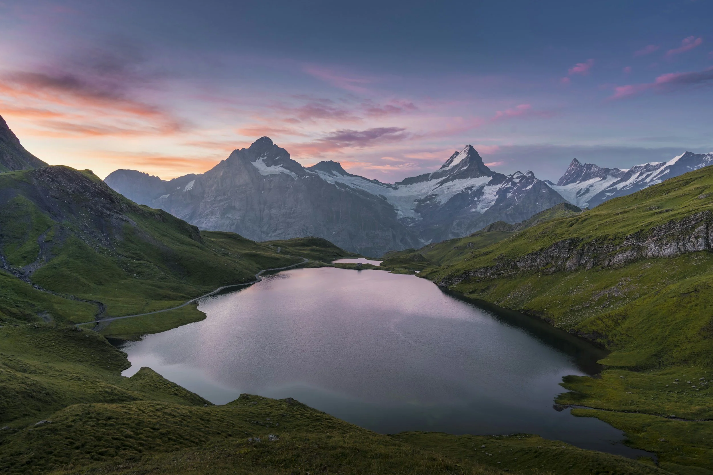 Snow-capped mountains at sunset with pink and purple clouds, green hills, and a calm lake in the foreground.