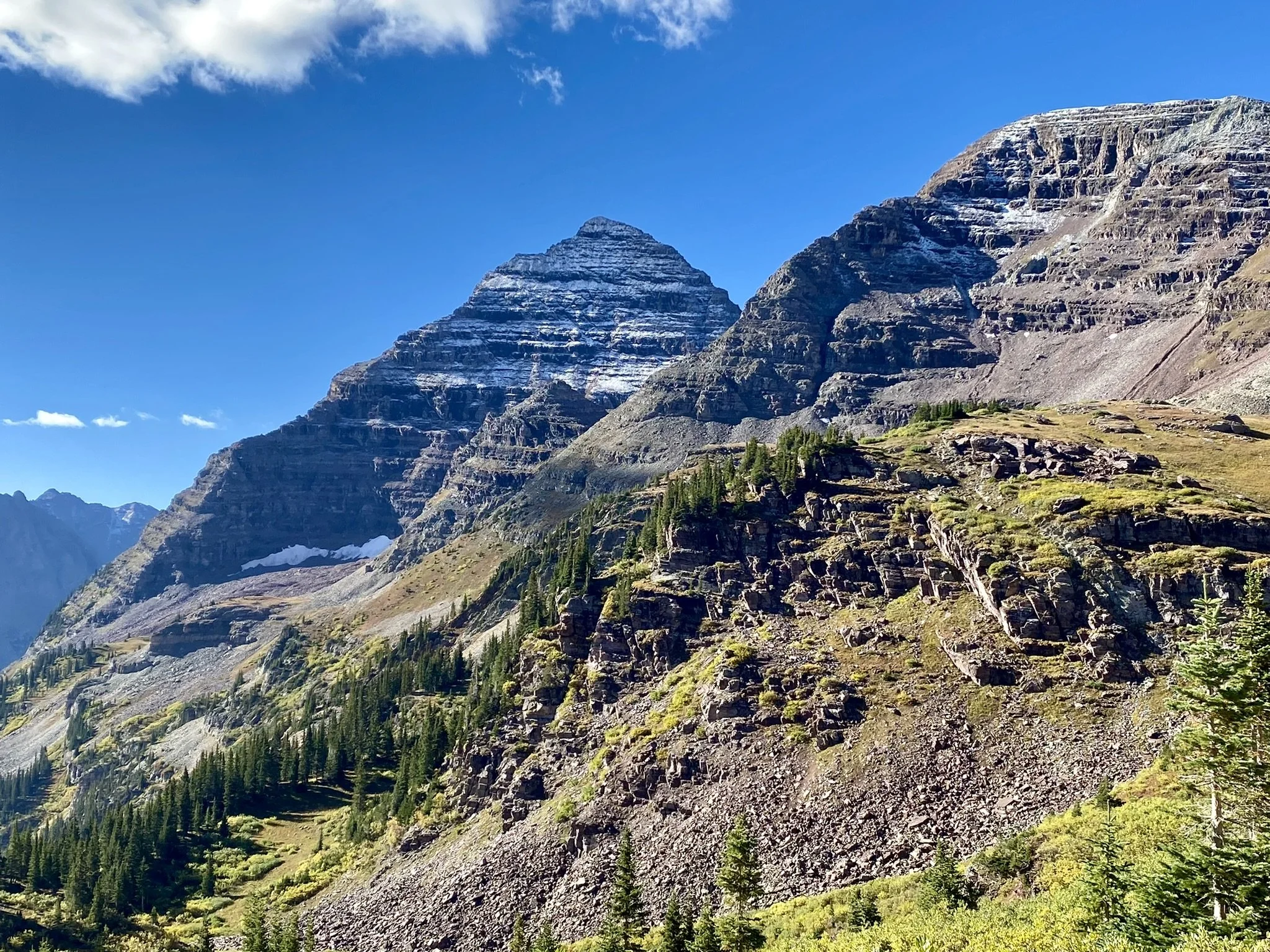 Majestic mountain range with layered rock formations, patches of snow, evergreen trees, and a bright blue sky with some clouds.