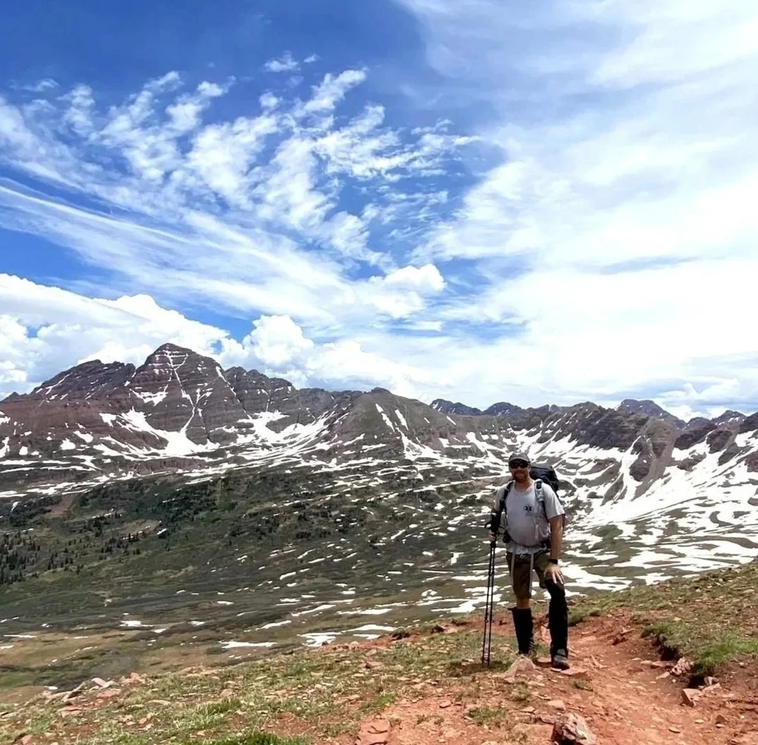 A man with hiking and backpacking gear standing on a trail with snow-capped Colorado Rocky mountains and a partly cloudy sky in the background.