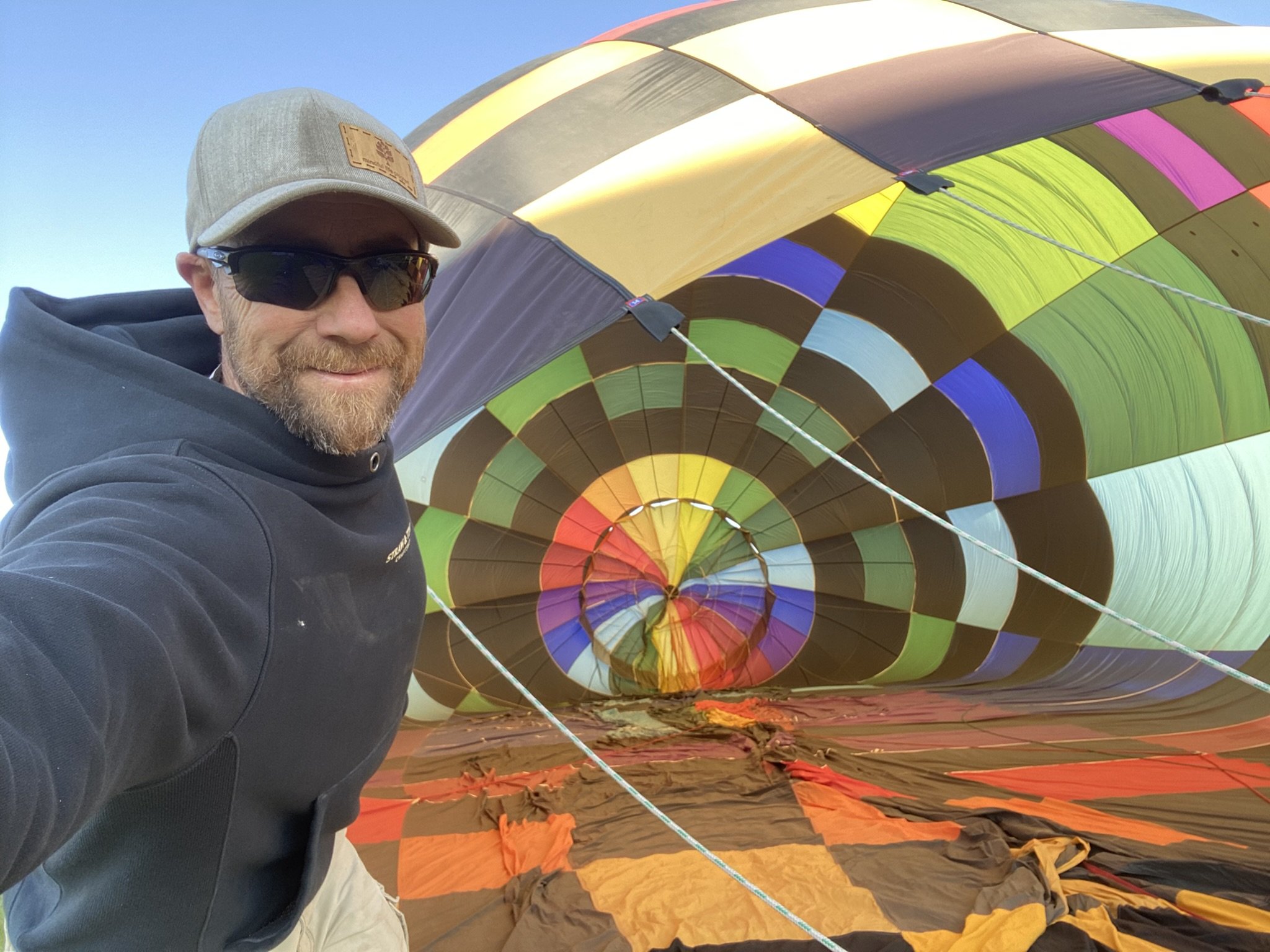 A man wearing sunglasses, a baseball cap, and a jacket, taking a selfie in front of a colorful hot air balloon that is lying on the ground.