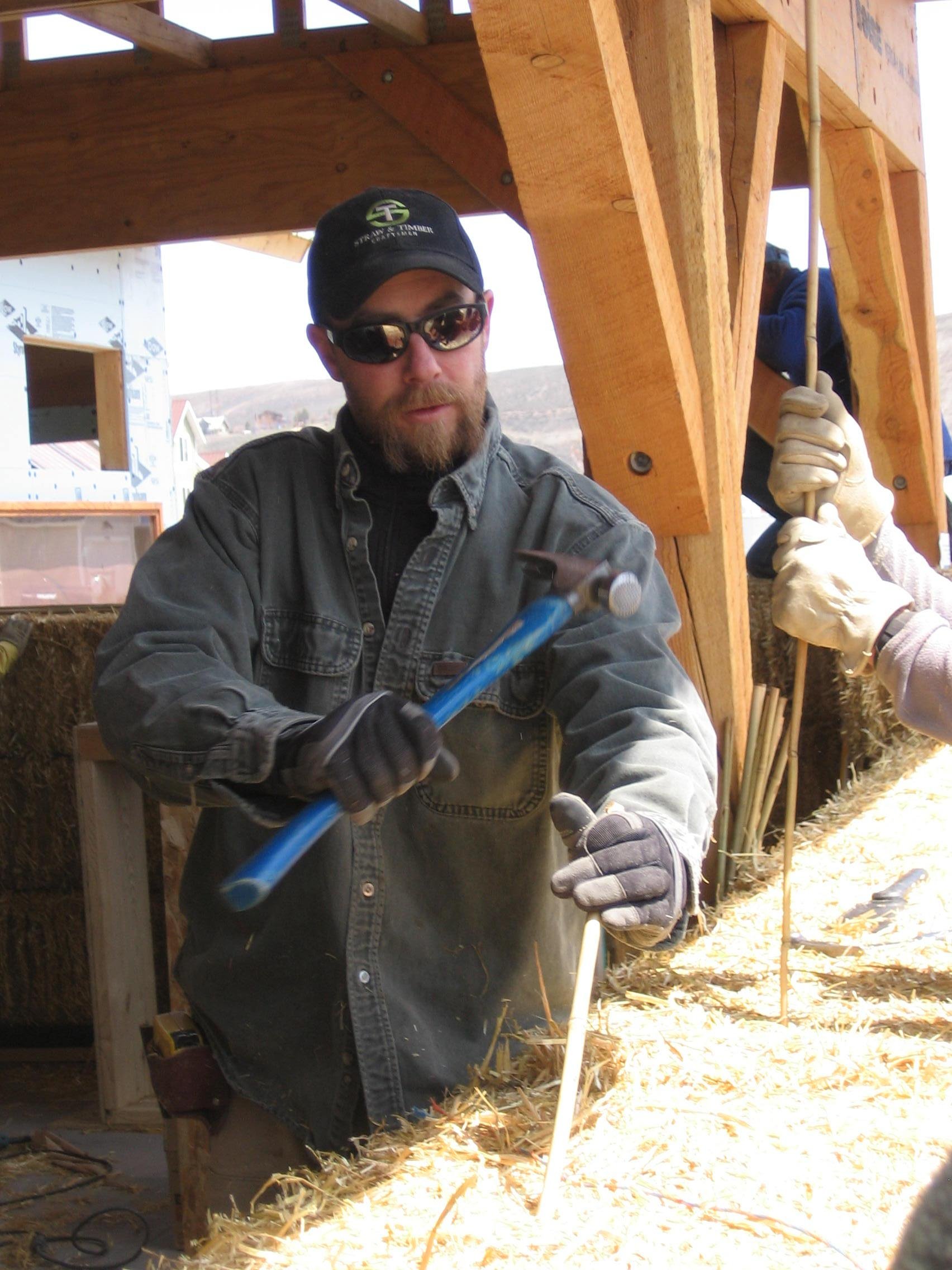 A man wearing sunglasses, gloves, and a denim jacket working on a construction site for a wooden structure, with wood framing and straw visible.