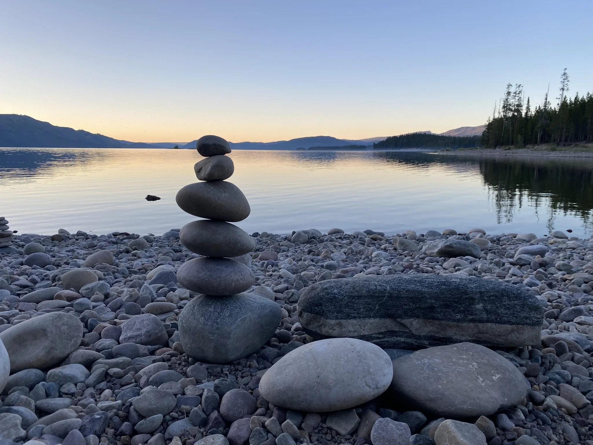 A stack of five smooth, rounded stones balanced on a rocky shore with a calm lake and forested hills in the background, during sunset or sunrise.