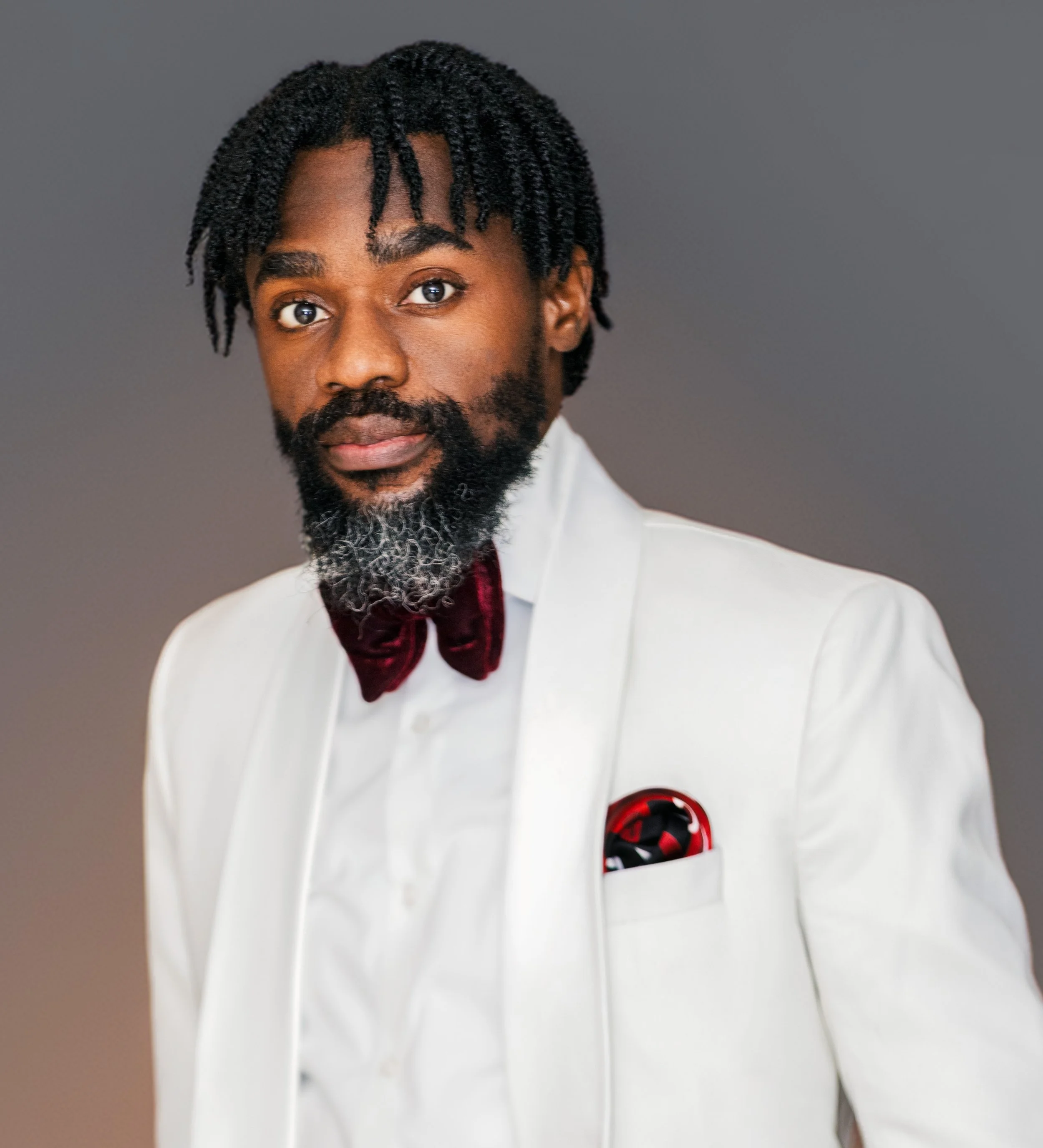 Portrait of a man with dark skin, beard, and braided hair, wearing a tan jacket over a graphic T-shirt, against a plain gray background.