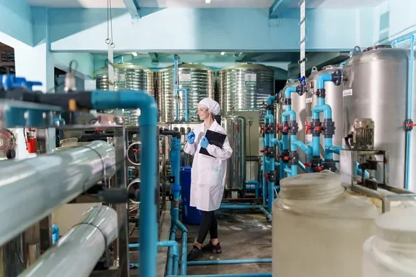 Vista general del interior de una industria en donde una mujer con el equipo correspondiente revisa los sistemas de tratamiento de agua.
