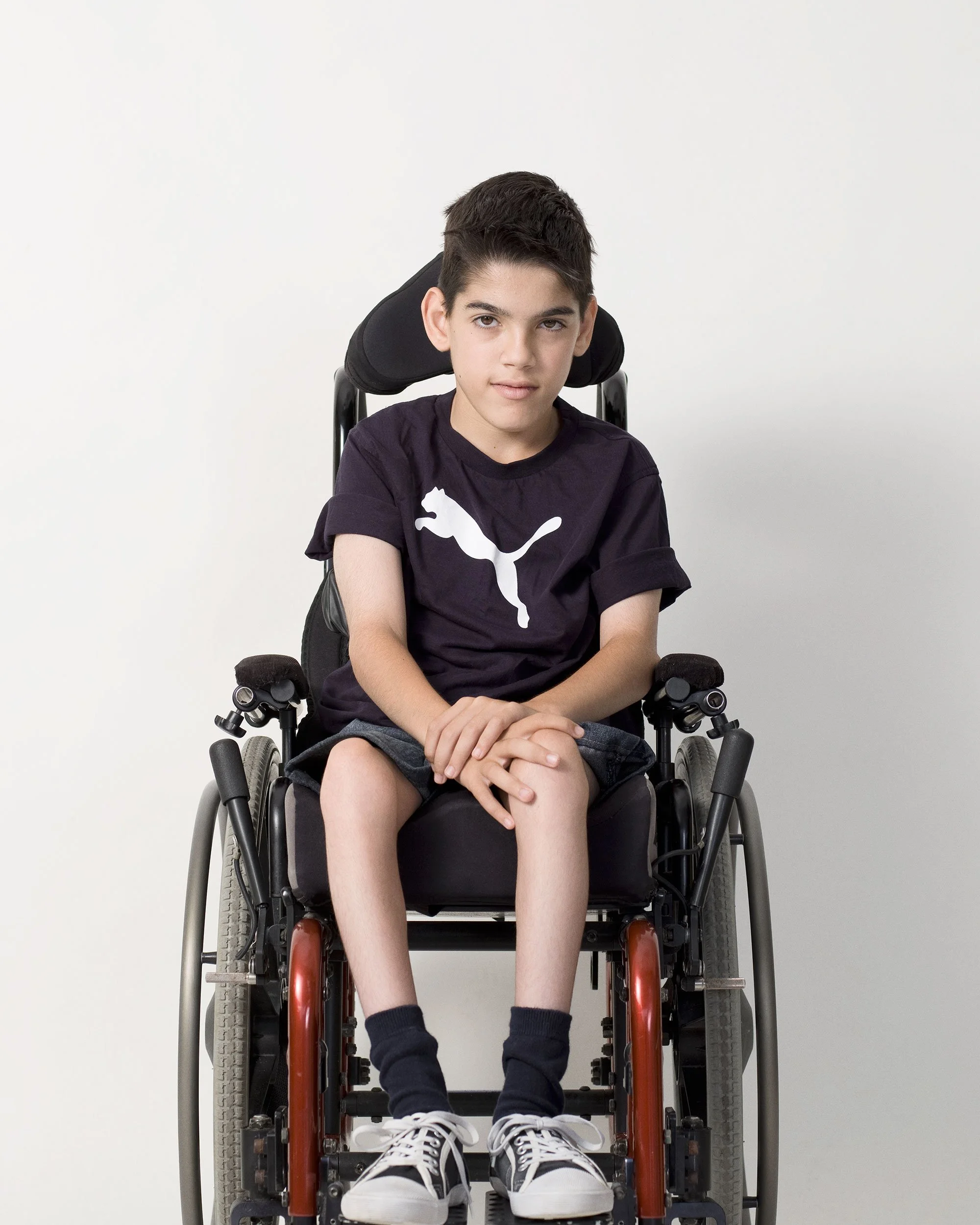 Portrait of a boy (Nicholas) with cerebral palsy sitting in a wheel chair with a Puma t-shirt and converse shoes by photographer David.A.Knight ©